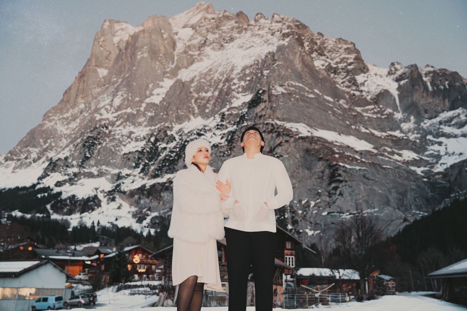 Couple holding hands in white winter outfits with snow-covered alpine village and mountain at golden hour.
