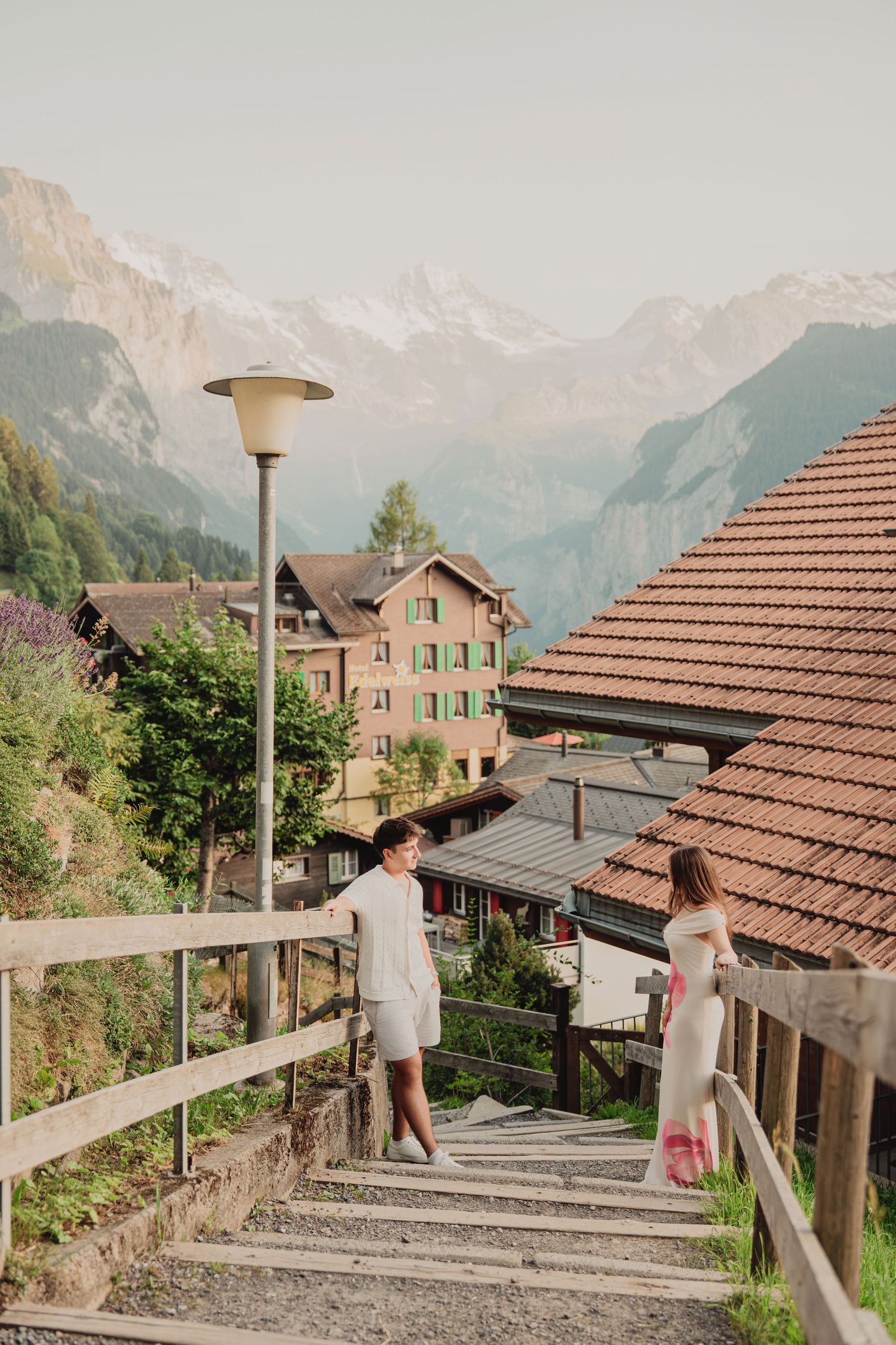 Couple on wooden staircase in picturesque Swiss village with snow-capped Alps and traditional chalets behind them.