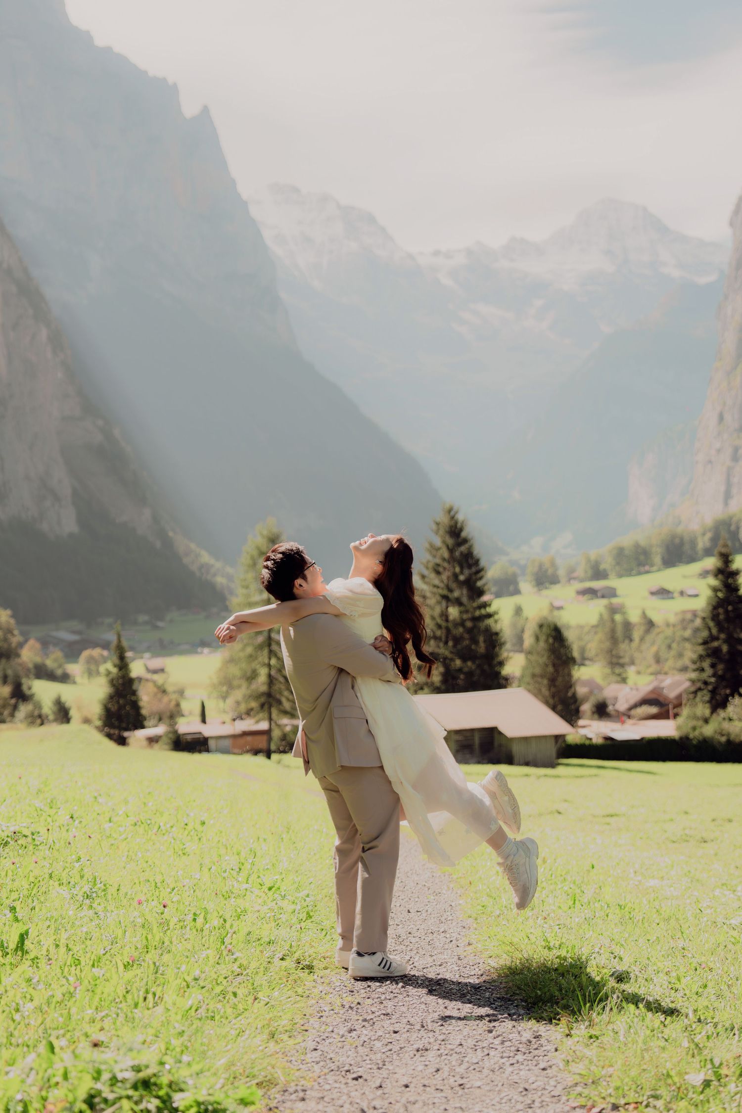 Groom lifting bride on a scenic Alpine valley path with mountains and green fields.