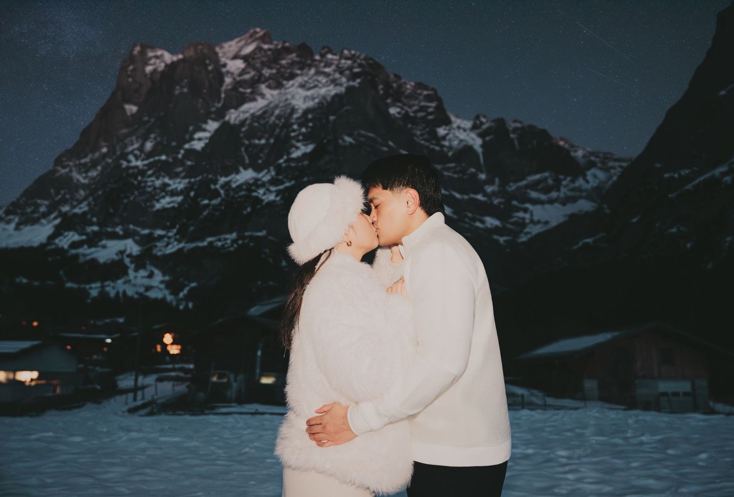 Couple sharing a kiss in white winter outfits under night sky with snow-covered mountain village behind.