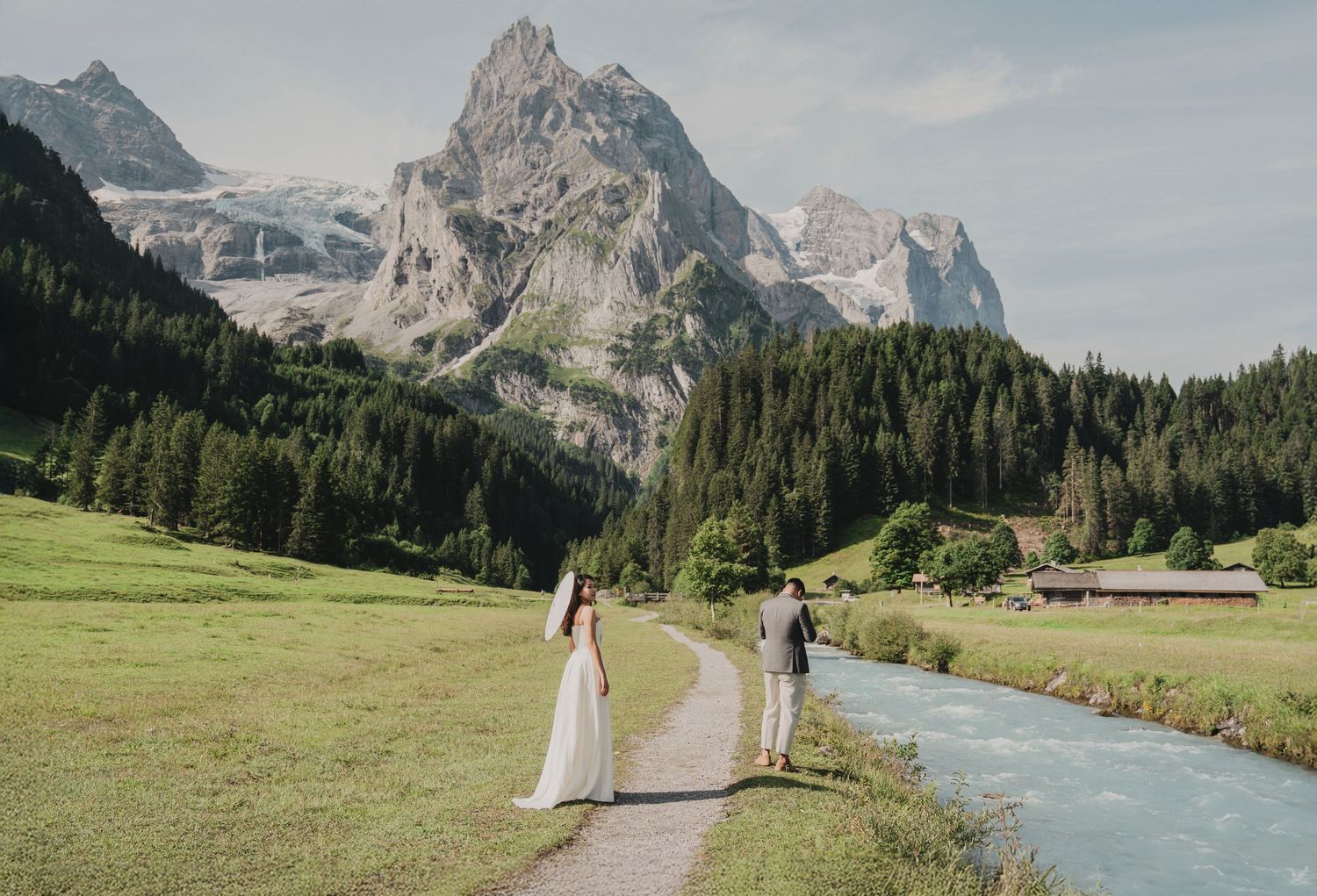 Wedding couple walks hand-in-hand along turquoise alpine river with stunning Swiss mountain peaks behind.