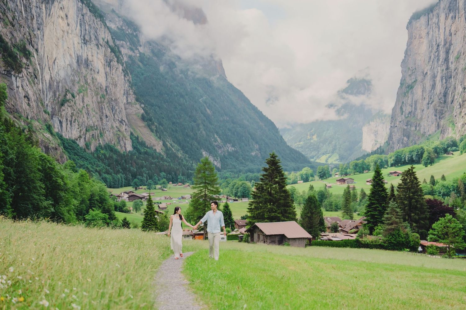 Couple walks hand in hand through lush green meadow path in scenic Lauterbrunnen valley with misty mountains.