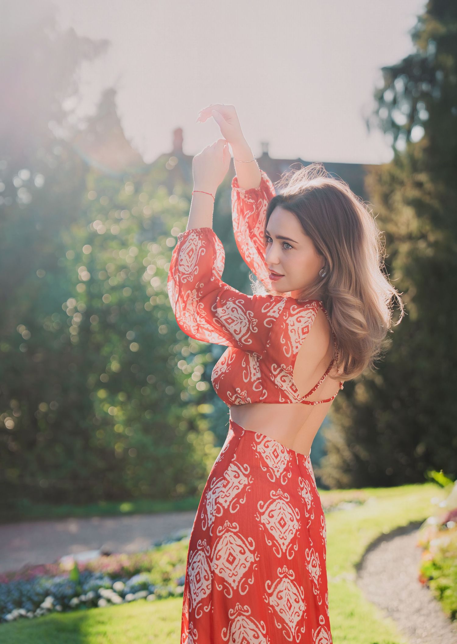Woman in backless red patterned dress posing in sunlit garden with arms raised.