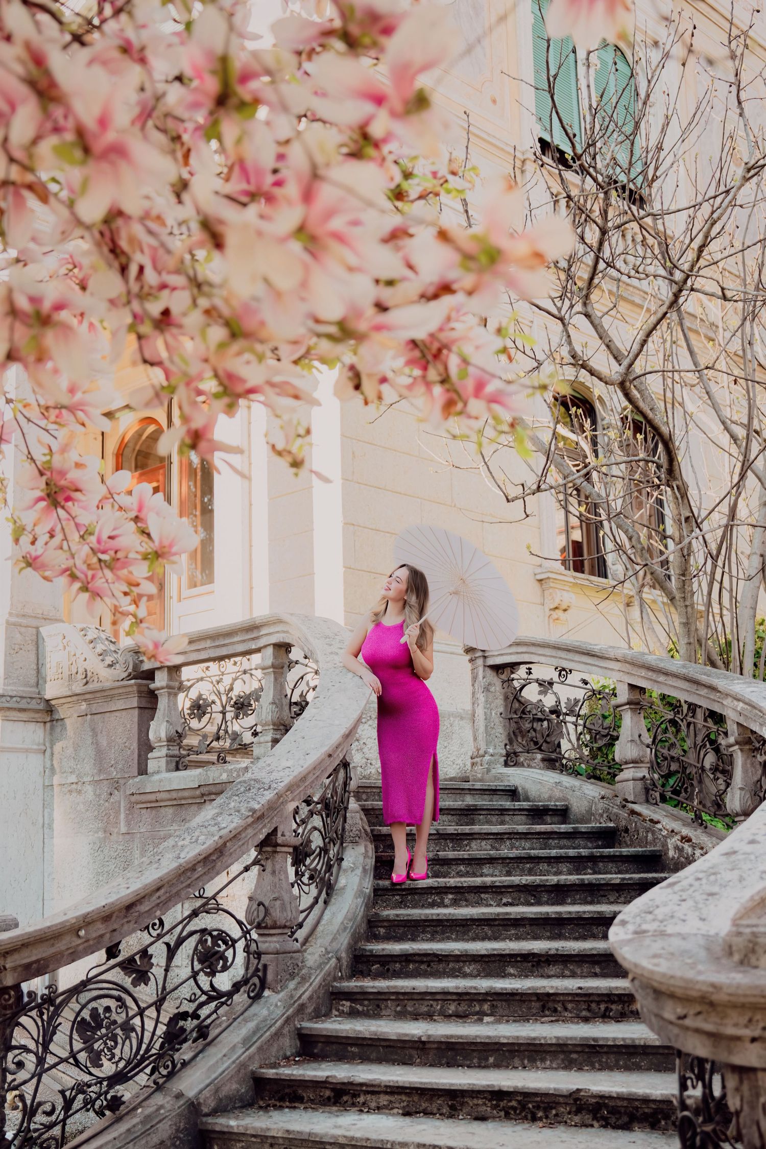 Woman in pink midi dress holding white parasol on curved stone staircase with cherry blossoms.