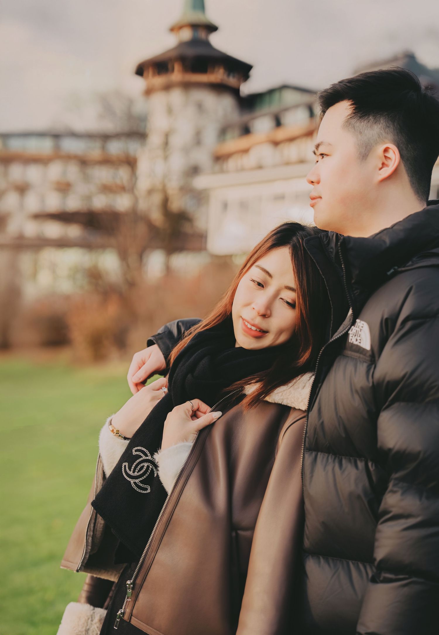 Asian couple standing close together outdoors with historic European castle tower in background.