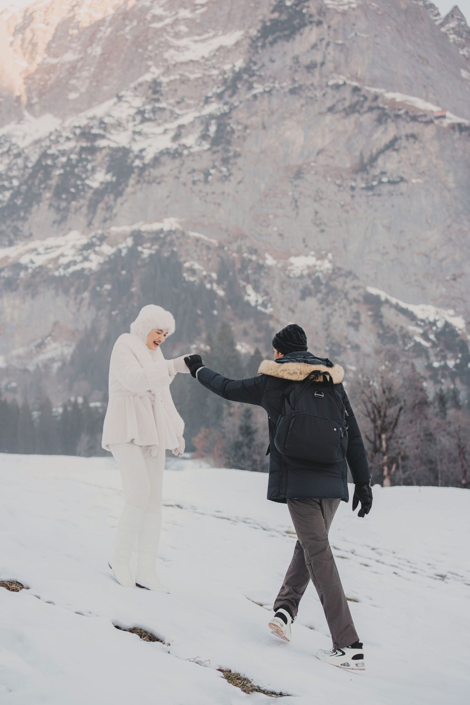 Couple holding hands walking in snowy alpine landscape with dramatic mountain backdrop in winter.