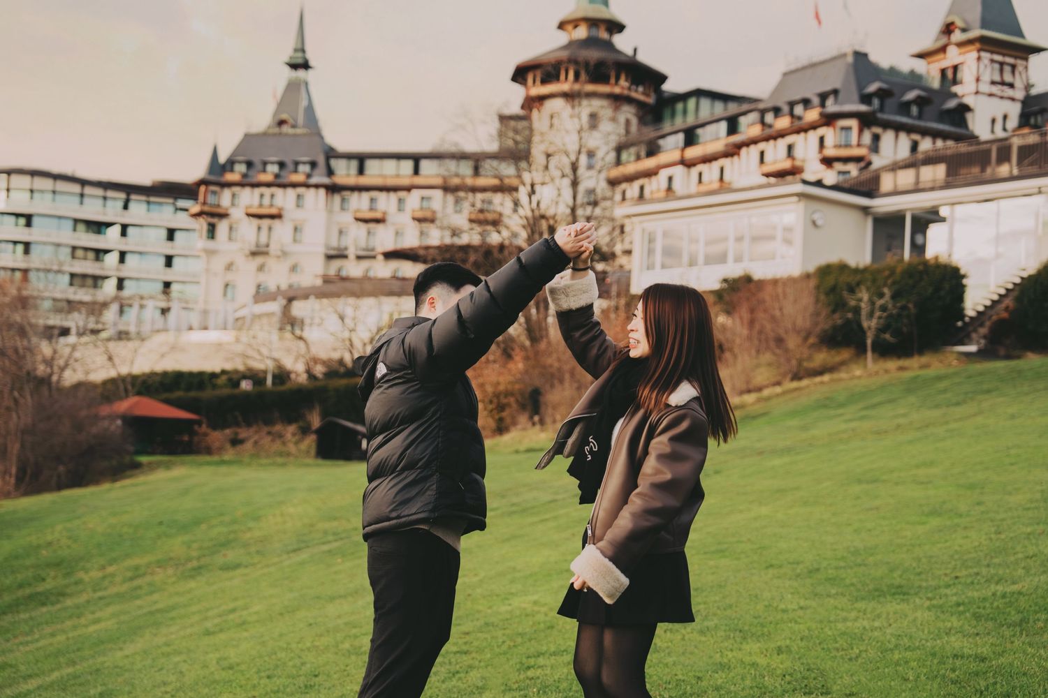 Couple dancing and holding hands on green lawn outside grand European castle.