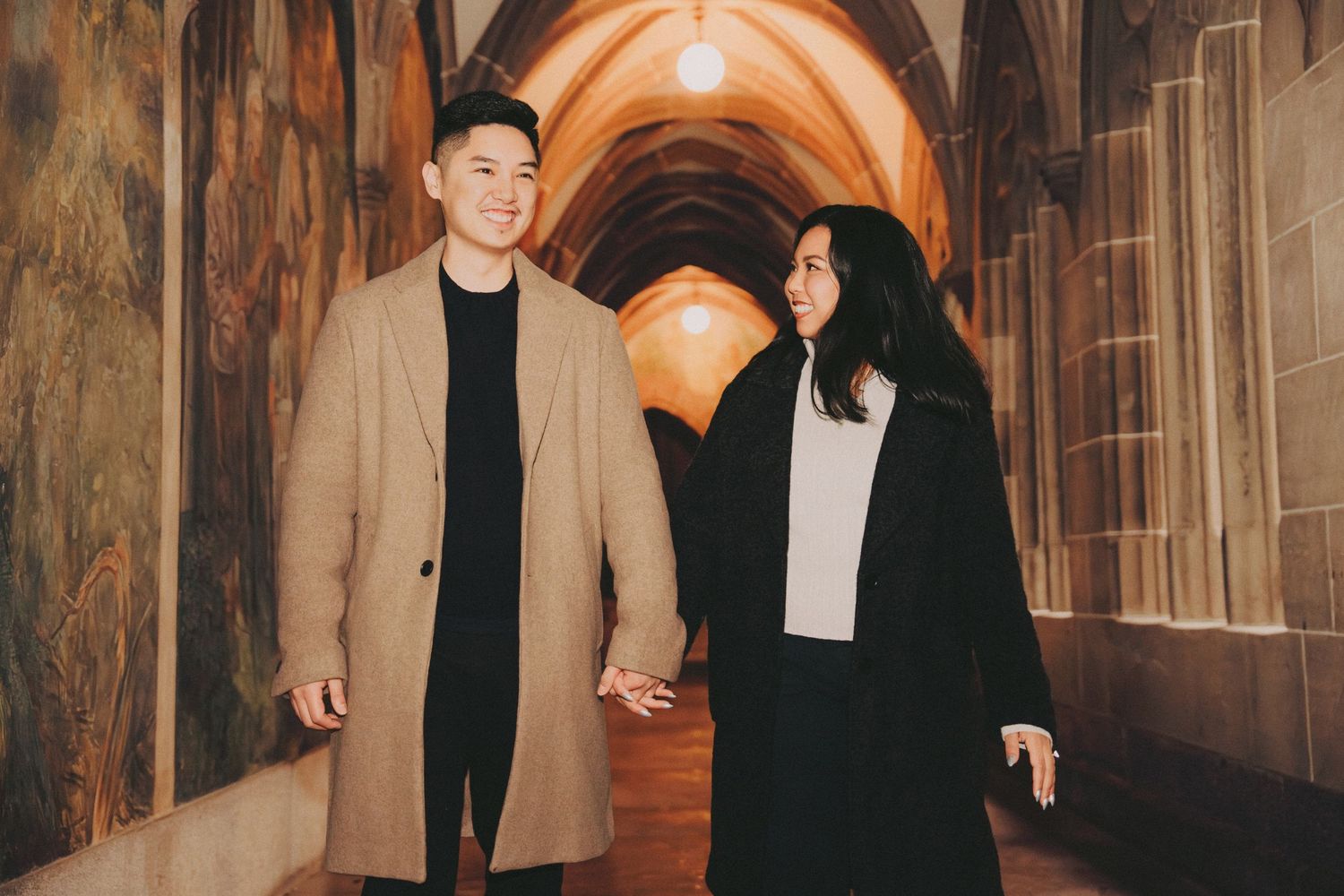 Couple holding hands walking through an arched stone corridor.