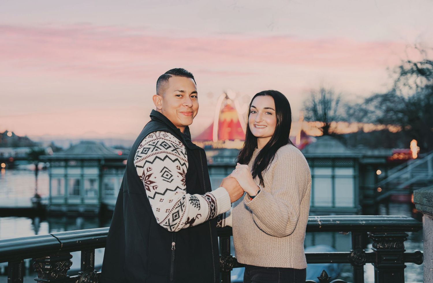 Couple holding hands at sunset with illuminated Zurich cityscape and river in background.