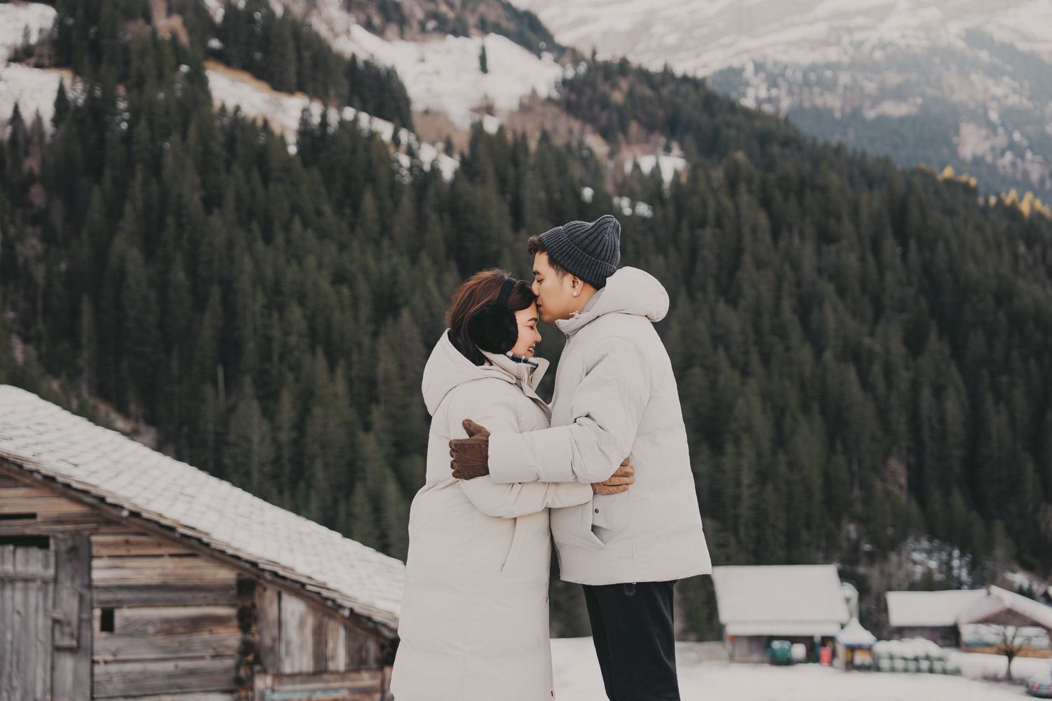 Couple sharing a tender kiss embrace in snowy alpine setting with wooden cabin and forest backdrop.