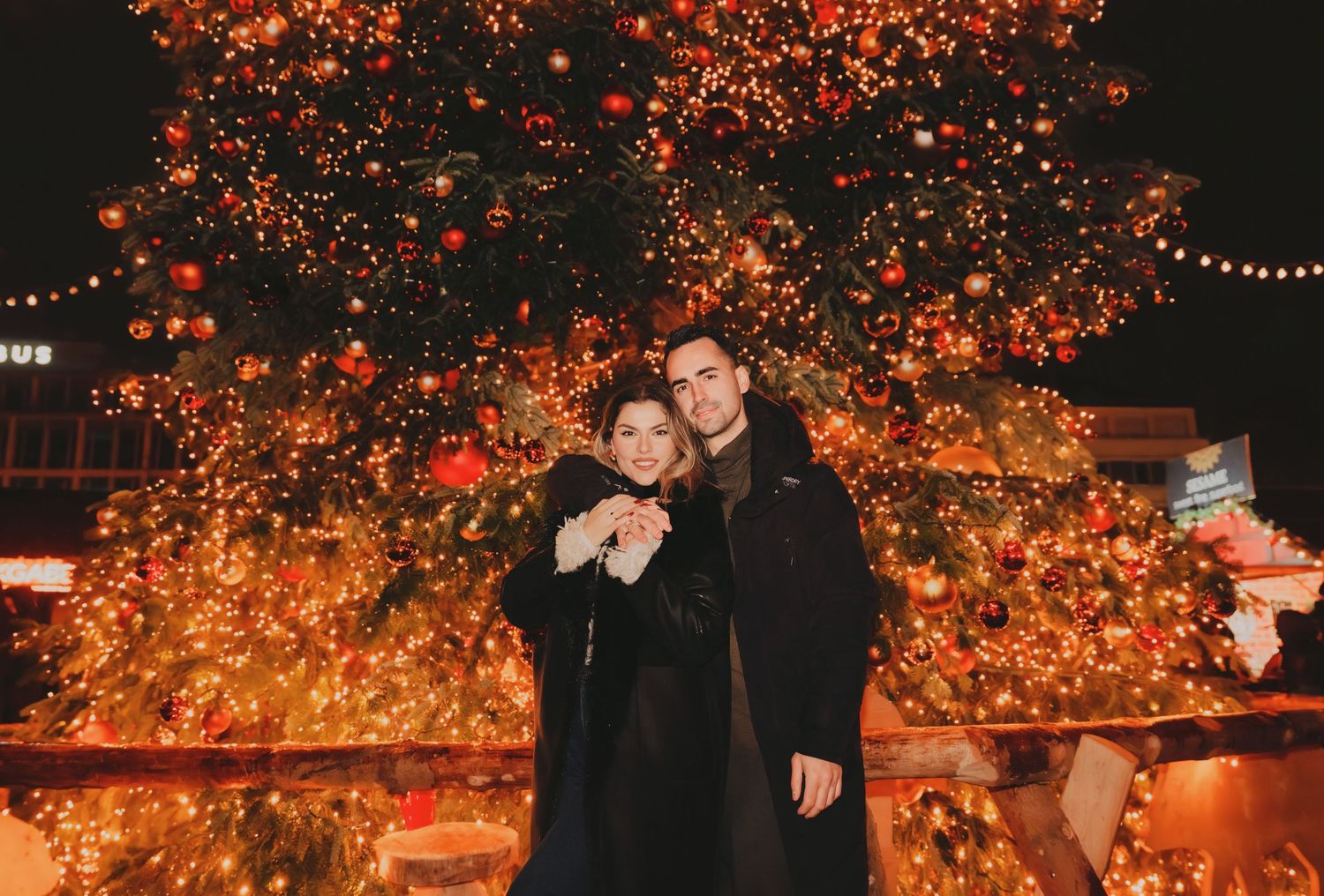 Couple smiling together in front of a beautifully lit Christmas tree at night.