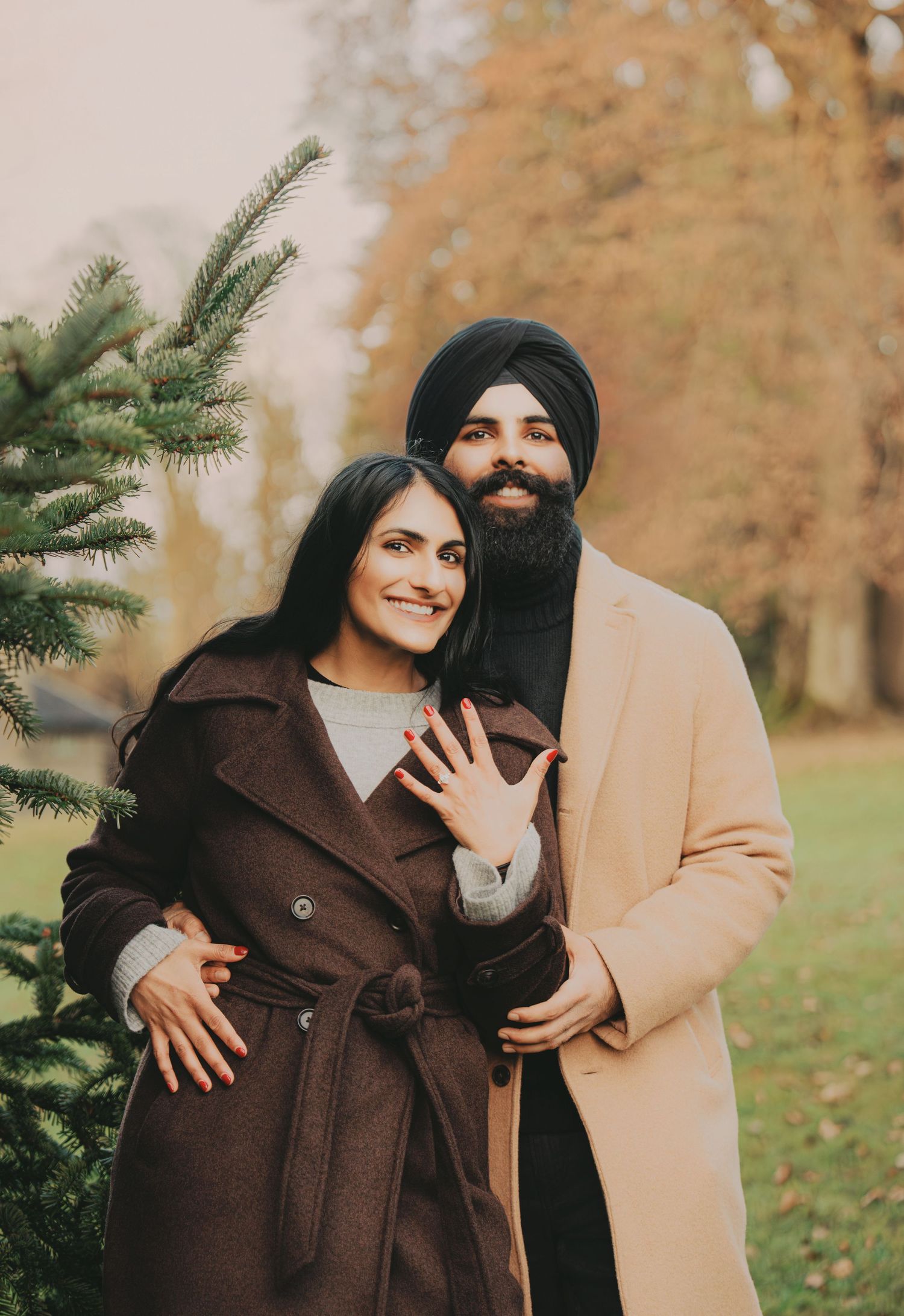 Smiling couple showing off engagement ring near evergreen tree in an autumn park setting.