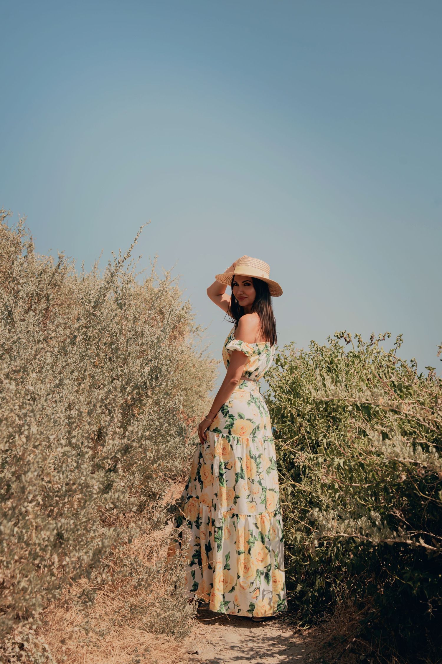 Woman in floral maxi dress and wide-brim straw hat posing on a dry Mediterranean nature path.