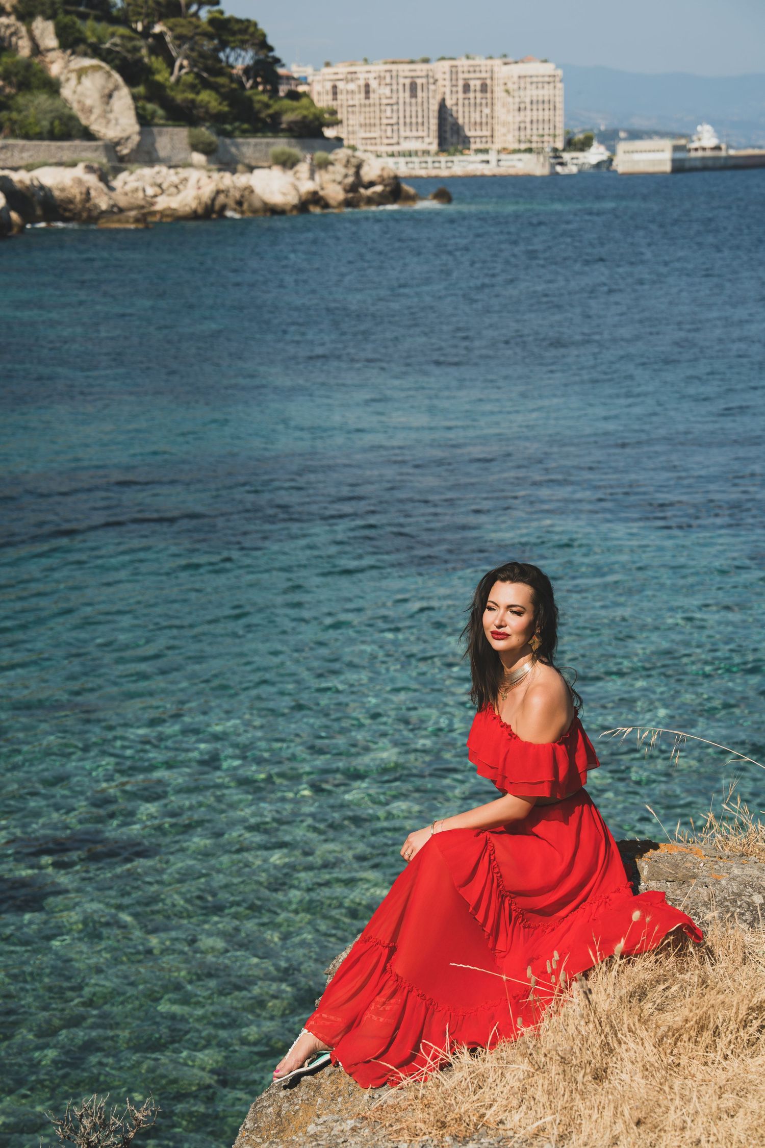 Woman in red off-shoulder dress sitting on coastal rocks with clear turquoise sea in background.