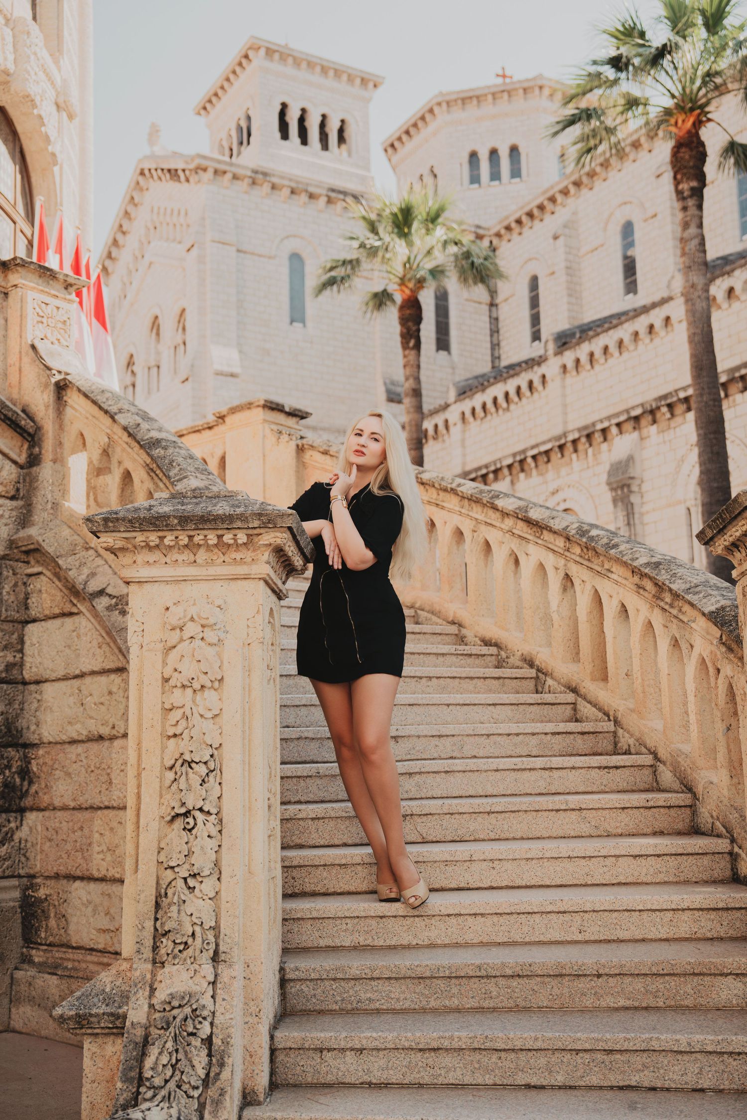 Elegant blonde woman in black dress smiling on decorative stone staircase with classical architecture and palm trees.