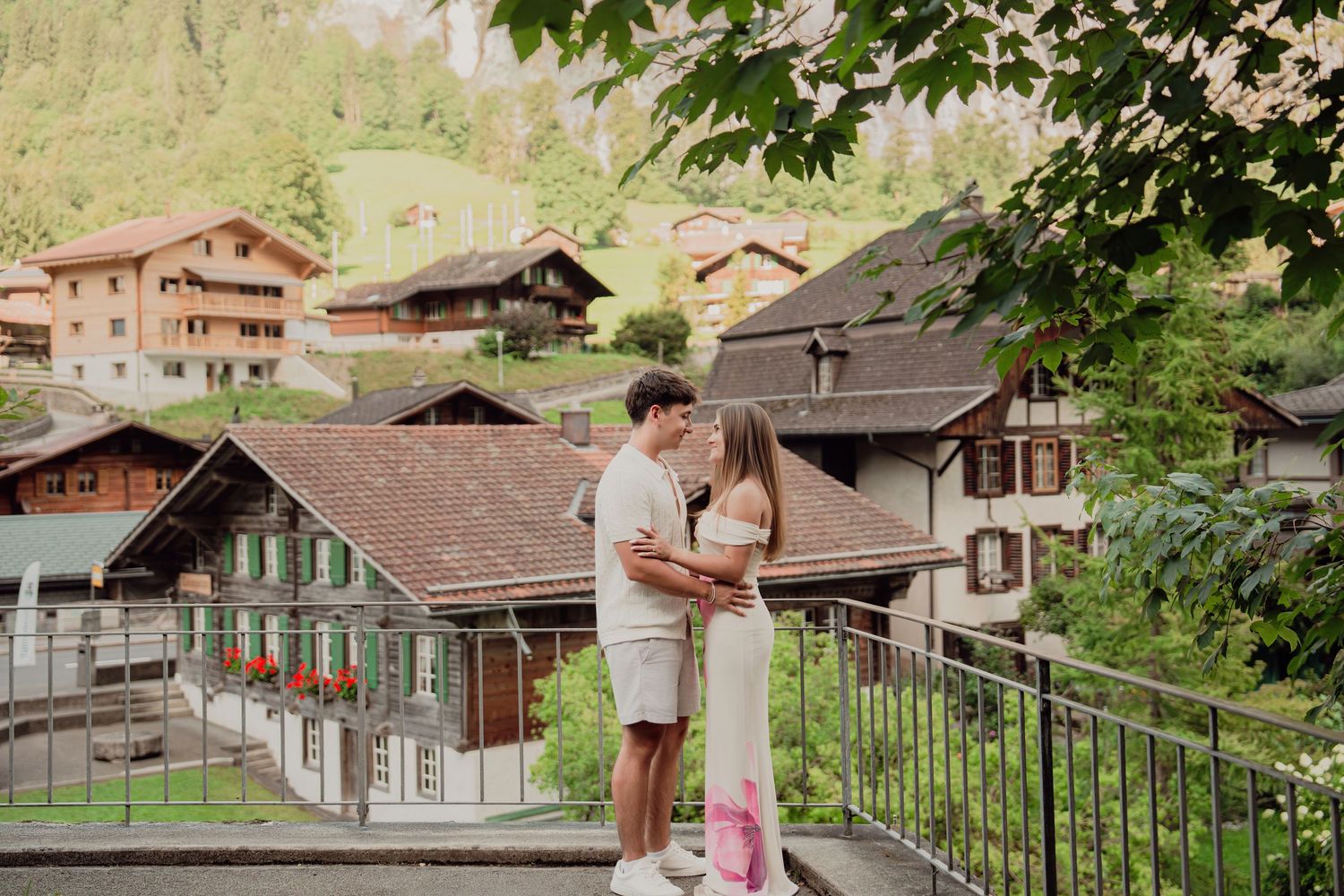 Young couple embracing on a balcony overlooking charming Swiss chalet homes in a mountain village.