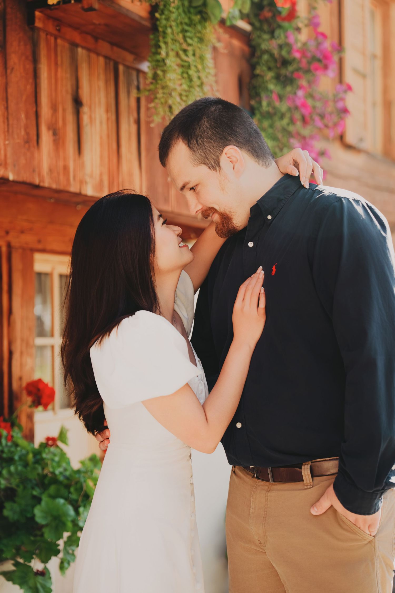 Couple sharing an intimate moment beside red geraniums and colorful flowers on a warm rustic wooden building facade.