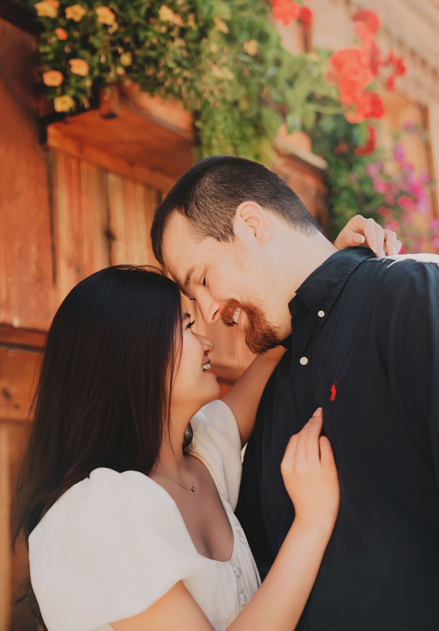 A couple sharing a kiss outdoors near a wooden wall adorned with vibrant red and orange flowers.