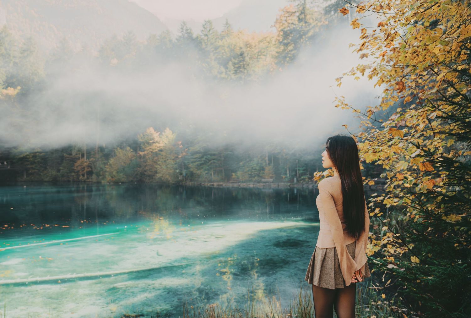 Young woman stands by a misty turquoise alpine lake surrounded by golden autumn trees and fog.