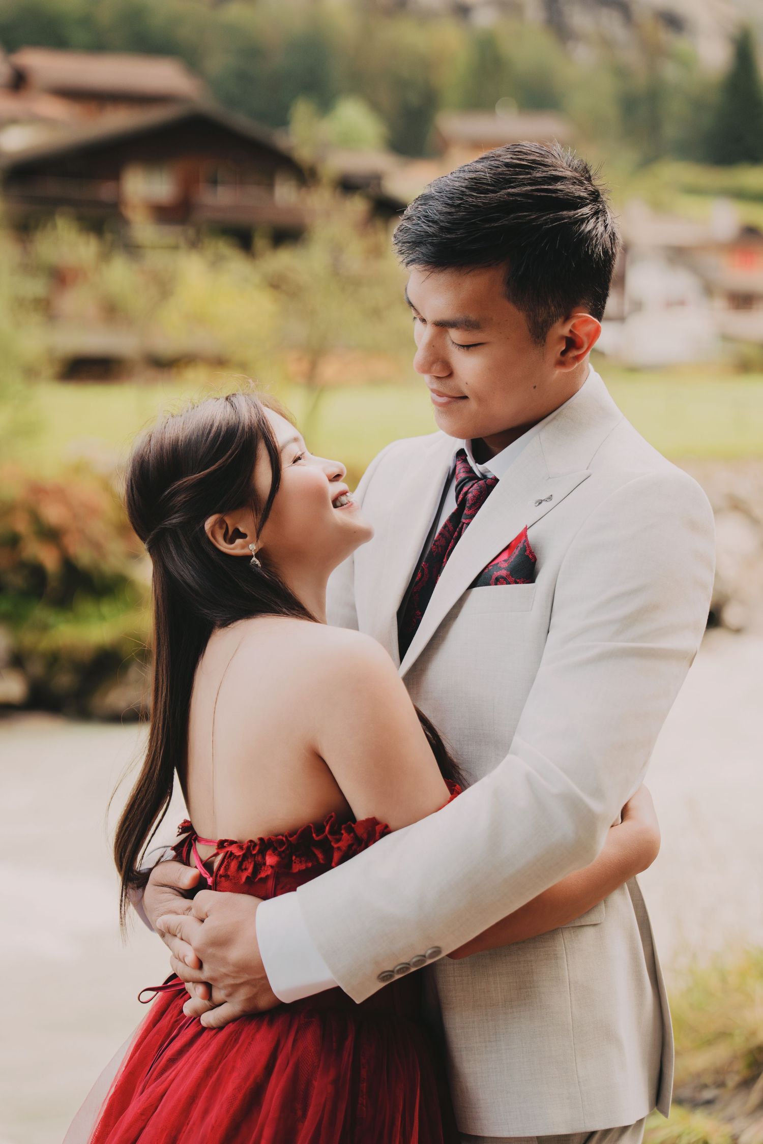 Joyful couple sharing a tender moment, bride in red strapless gown and groom in light gray suit outdoors.