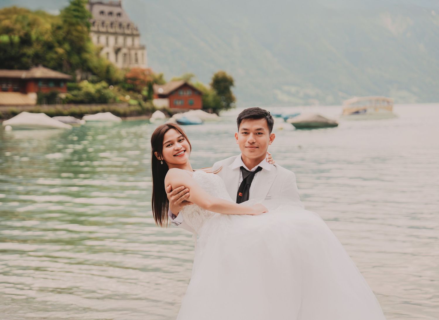 Groom carrying bride in white gown by a serene alpine lake with lush green mountains behind them.