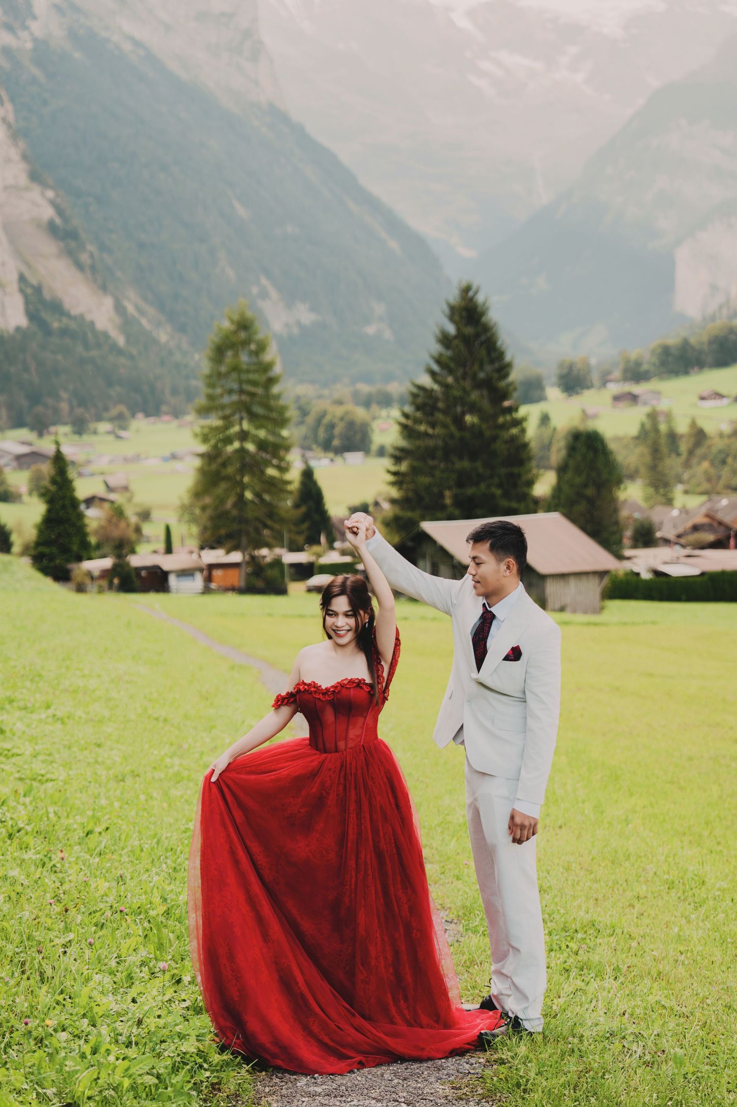 Couple dancing on alpine path, bride in red off-shoulder gown, groom in white suit, Switzerland.