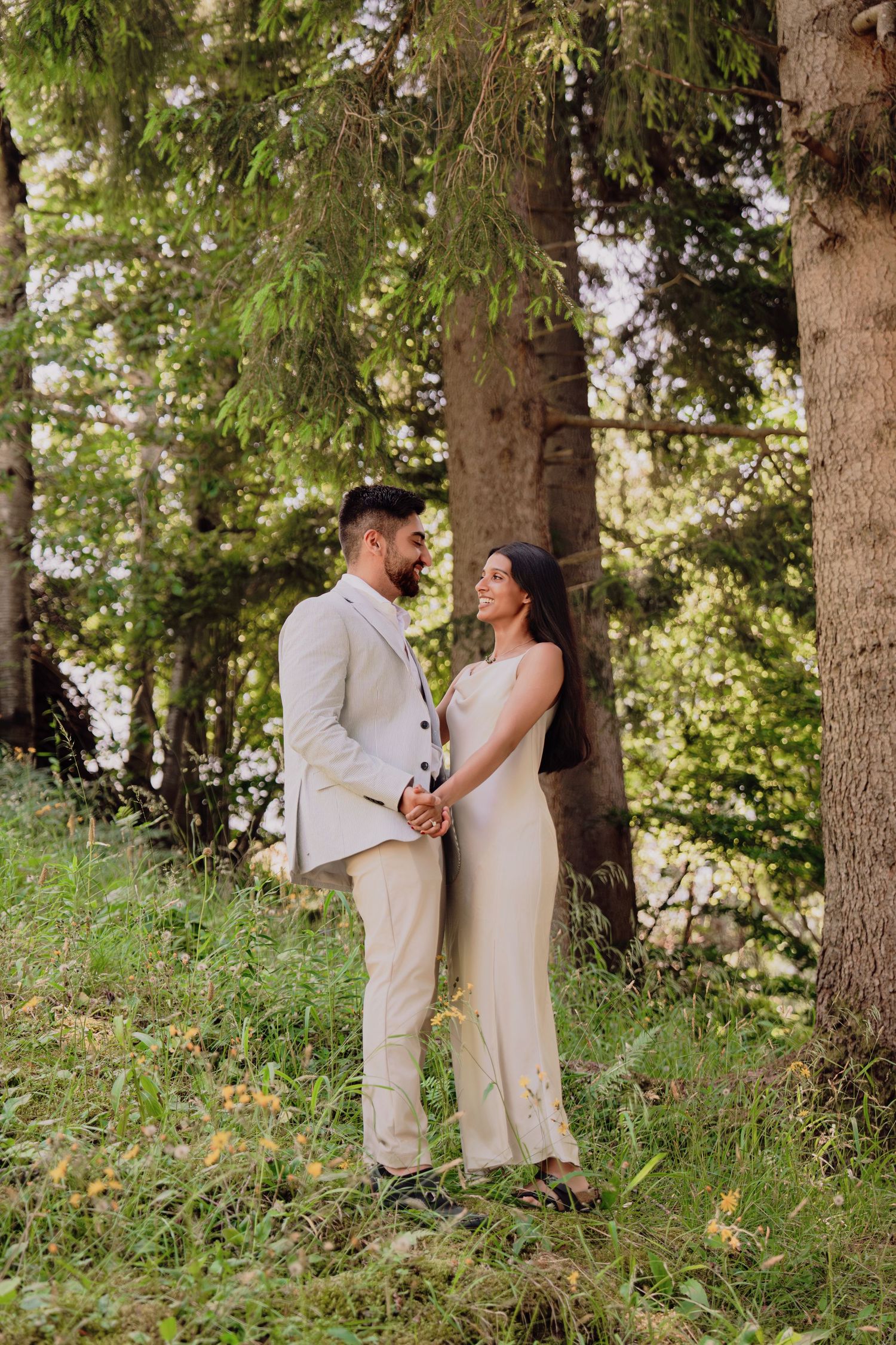 Smiling couple holding hands while standing among tall pine trees in a lush green forest setting.