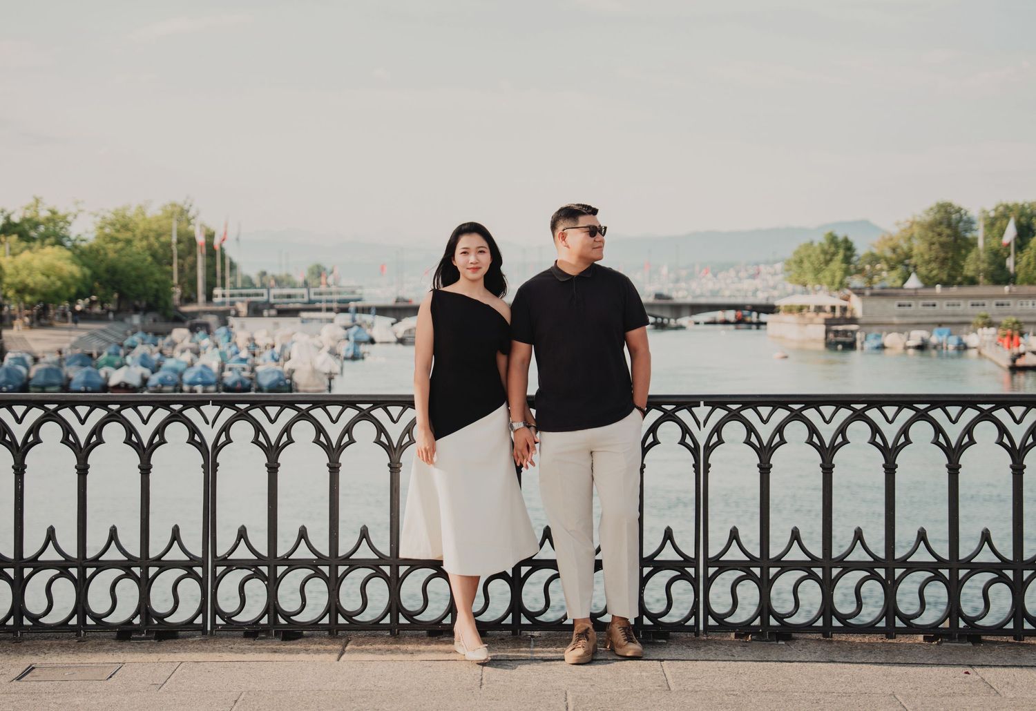 Couple standing together by decorative iron fence with Zurich lake harbor and boats behind them.