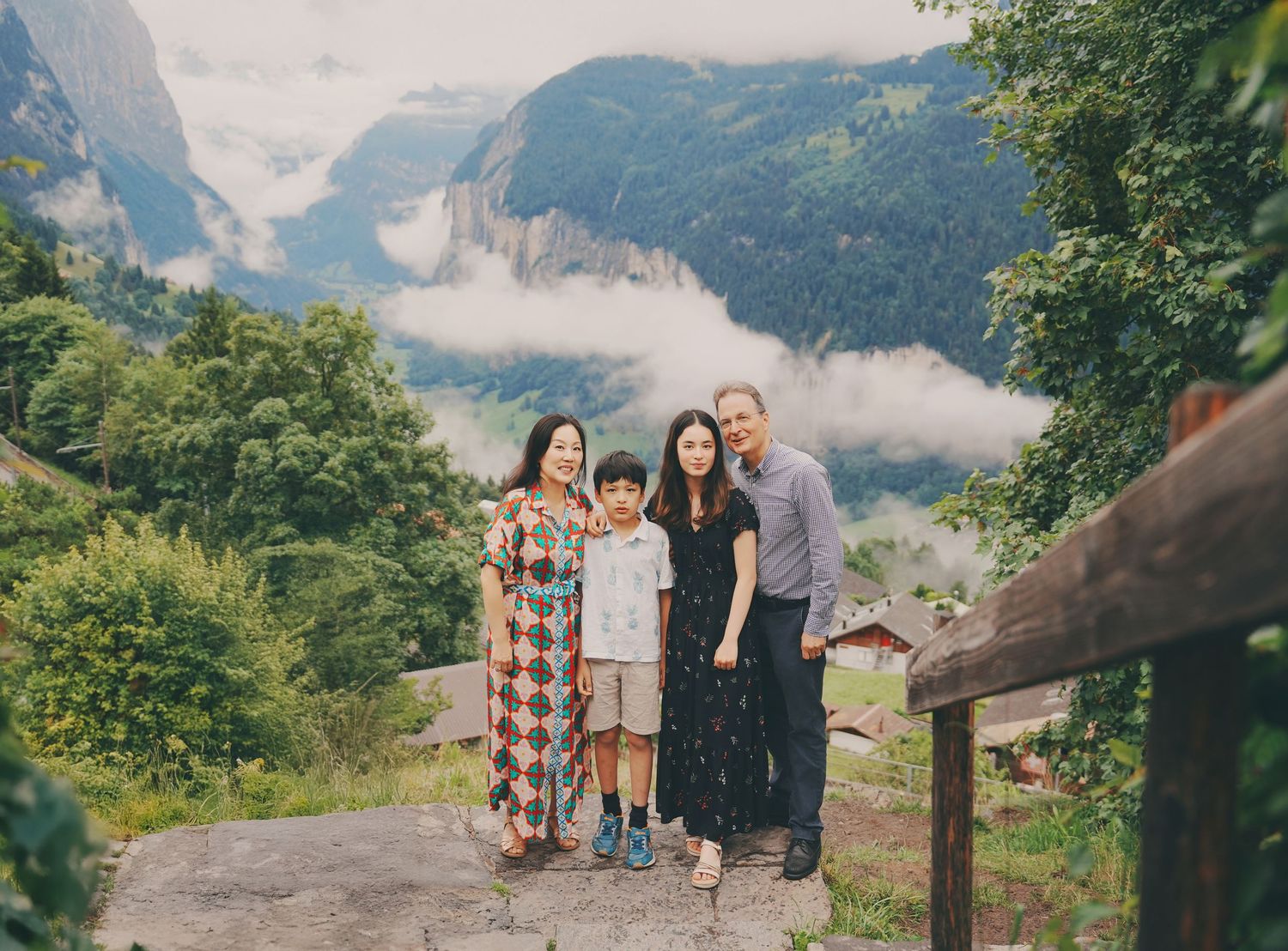 Asian family of four smiling together on stone overlook with misty Swiss Alpine valley and mountains behind them.