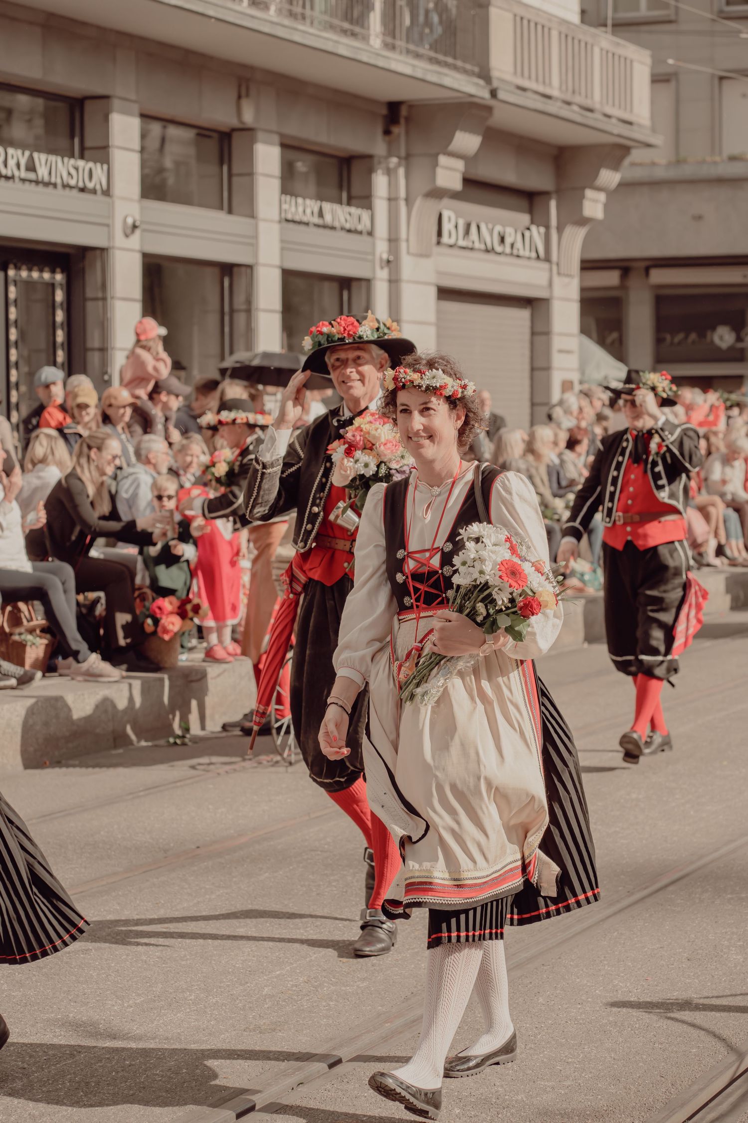 Woman in traditional Swiss folk costume with flower crown carrying bouquet during a sunny street parade near luxury shops.