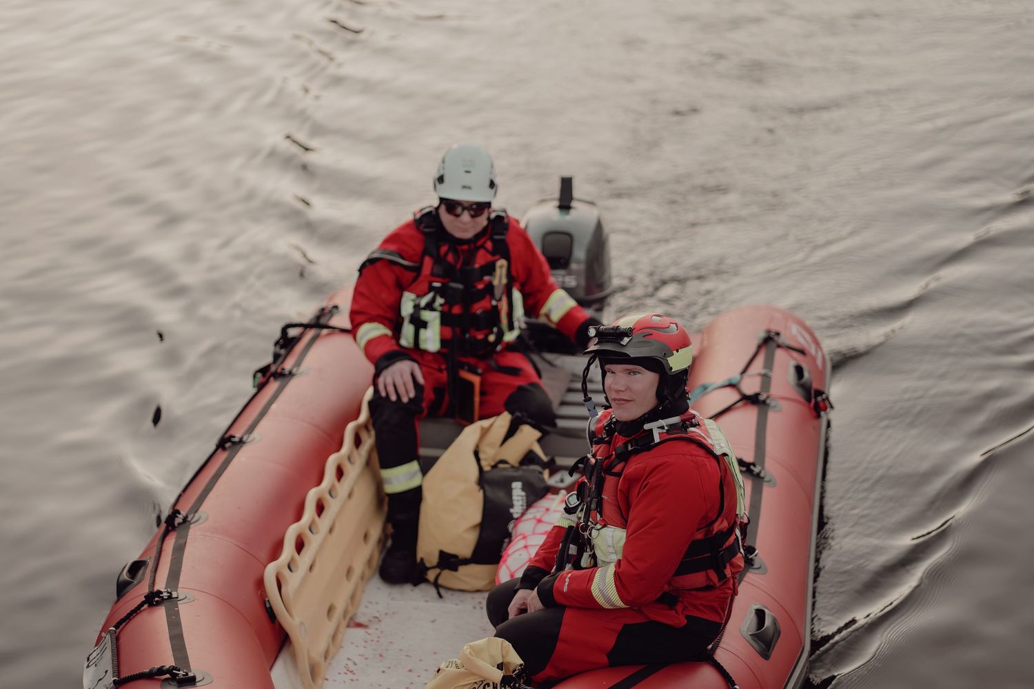 Two emergency rescue personnel in red gear navigating an inflatable boat across calm grey water.