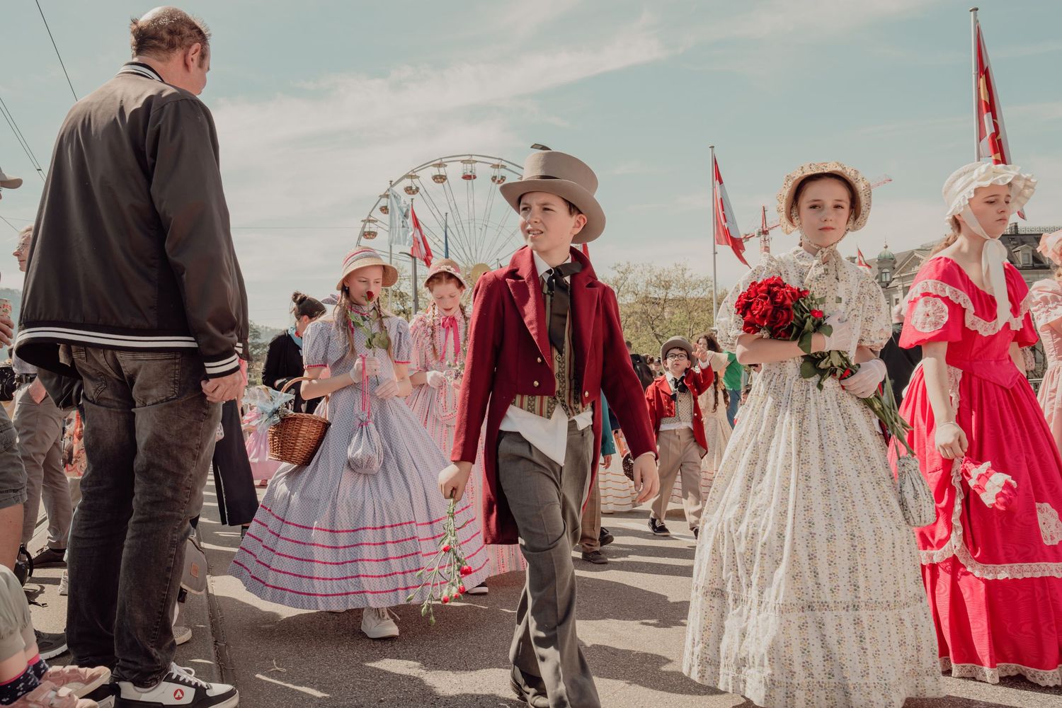 Children and adults in Victorian-era costumes parade at a festive outdoor event with a Ferris wheel in the background.