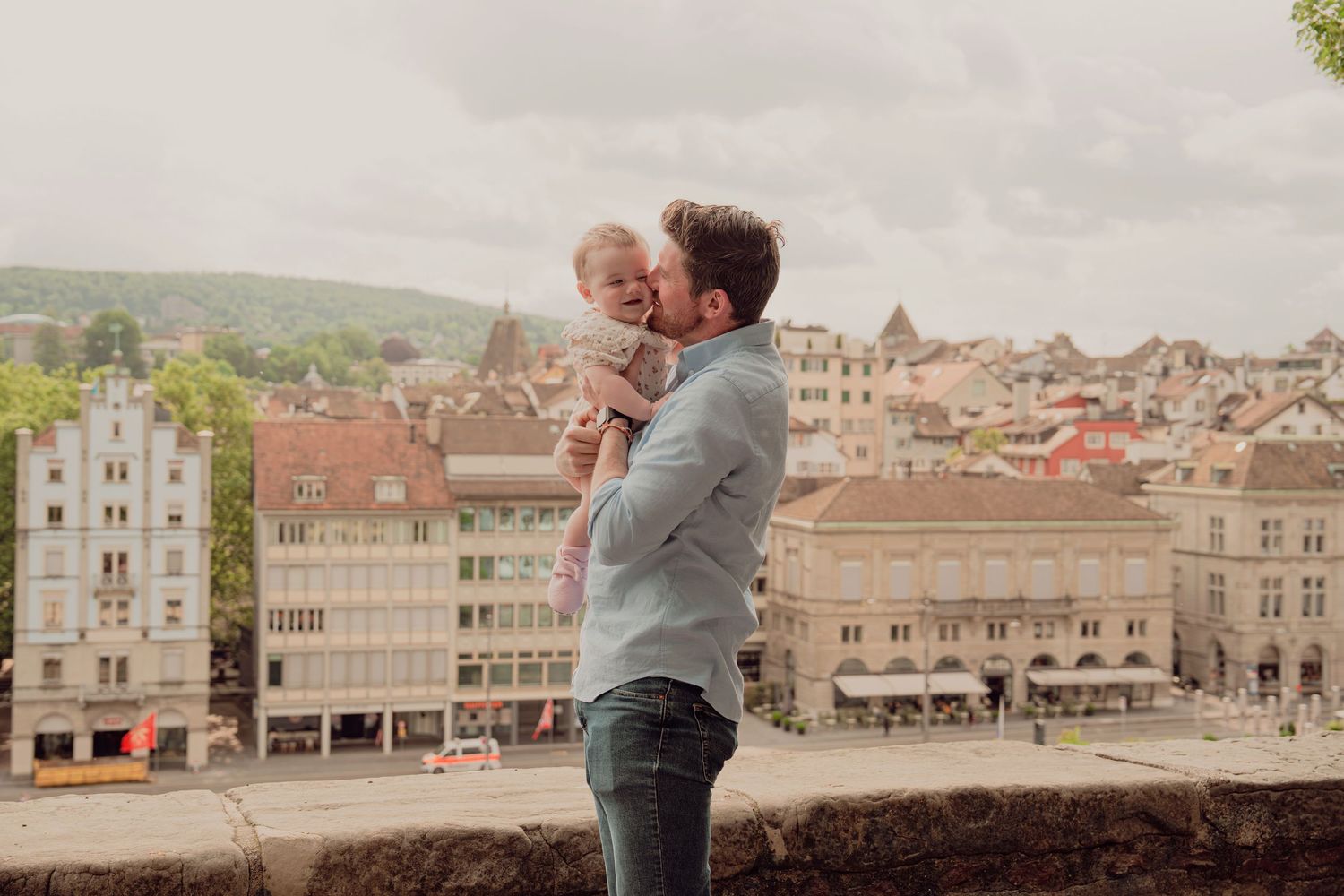 Father tenderly kissing baby while standing on overlook above European old town.