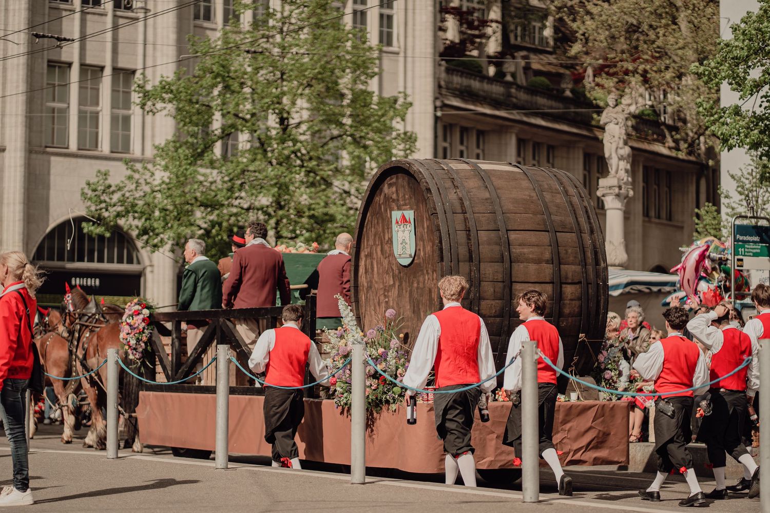 People in traditional attire push a large wooden barrel float through city streets during a festive parade.