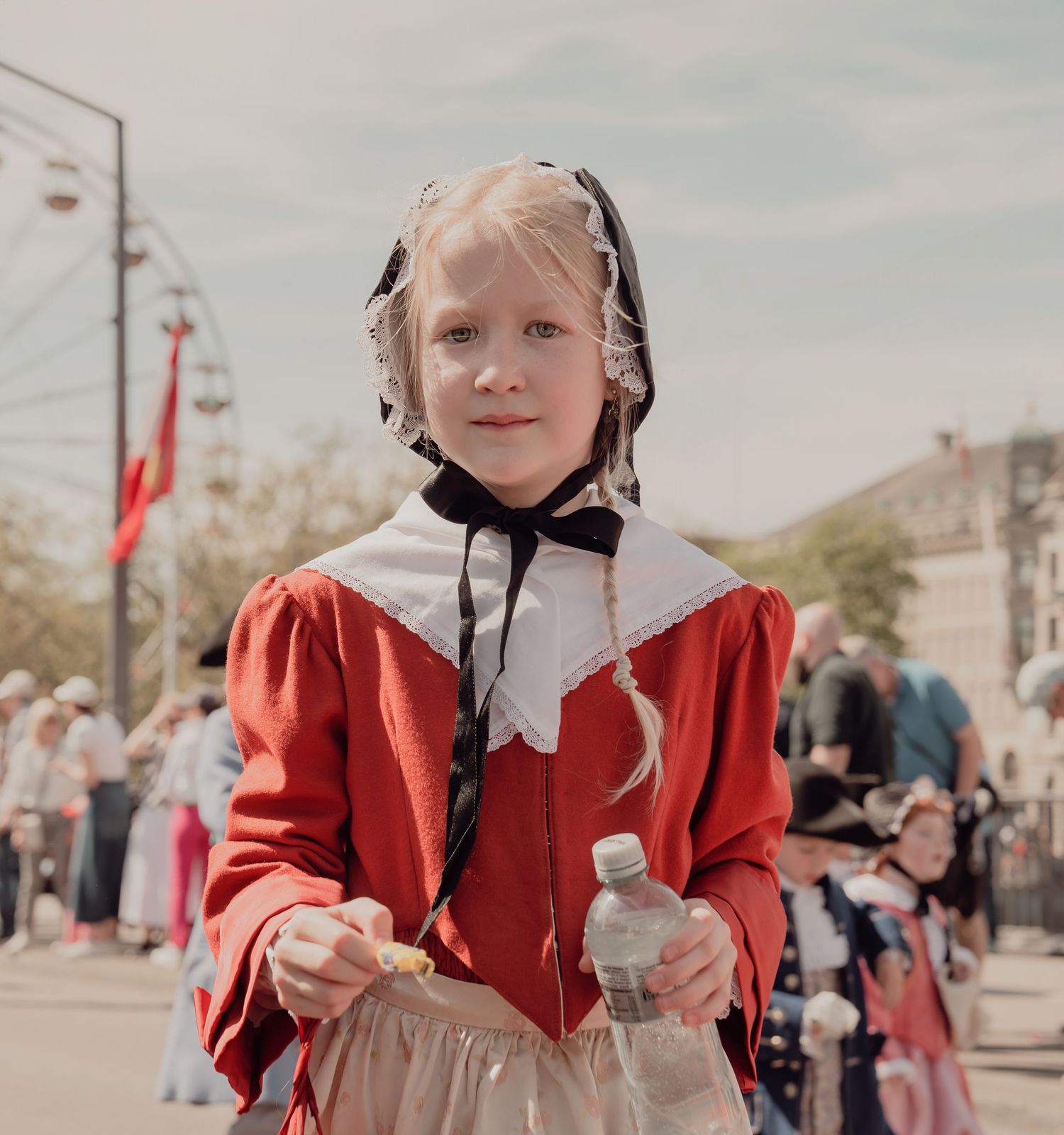 Young girl in traditional red costume with white bonnet at a festive outdoor parade.