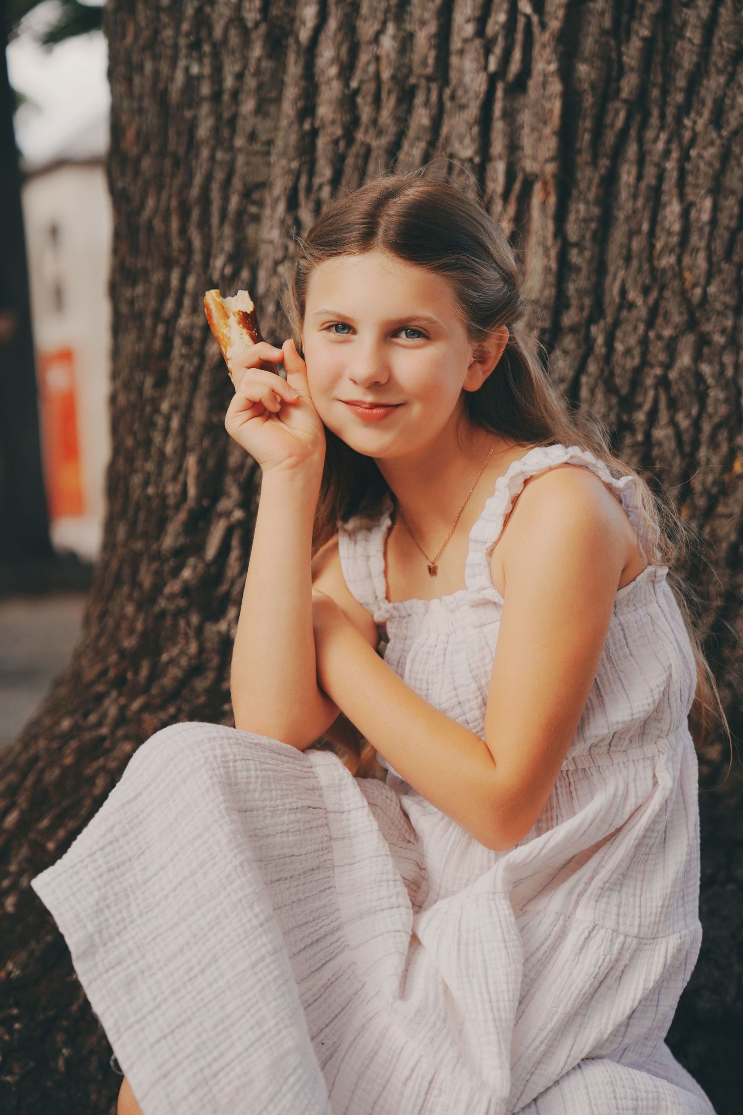 Smiling preteen girl in white dress sitting against a tree holding a treat.