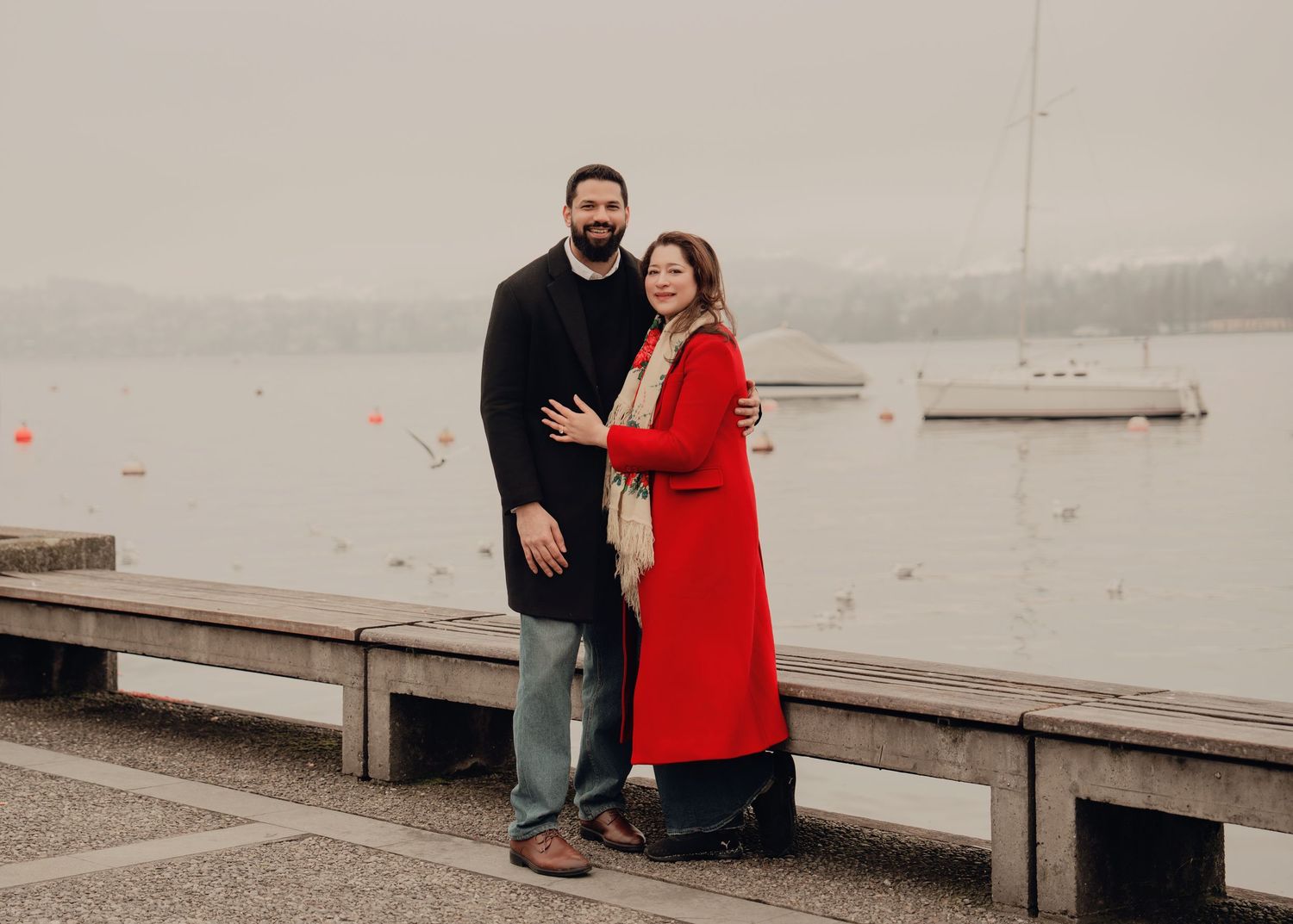 Couple embracing on misty lakeside promenade, man in black coat and woman in red long coat.