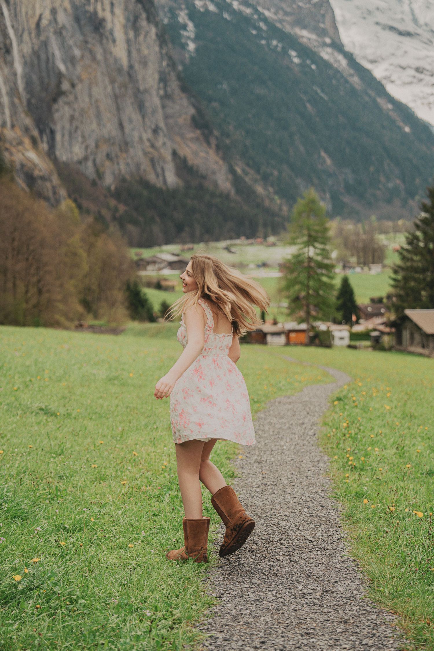 Blonde woman spinning in pink floral dress and brown boots on scenic alpine trail in Switzerland.