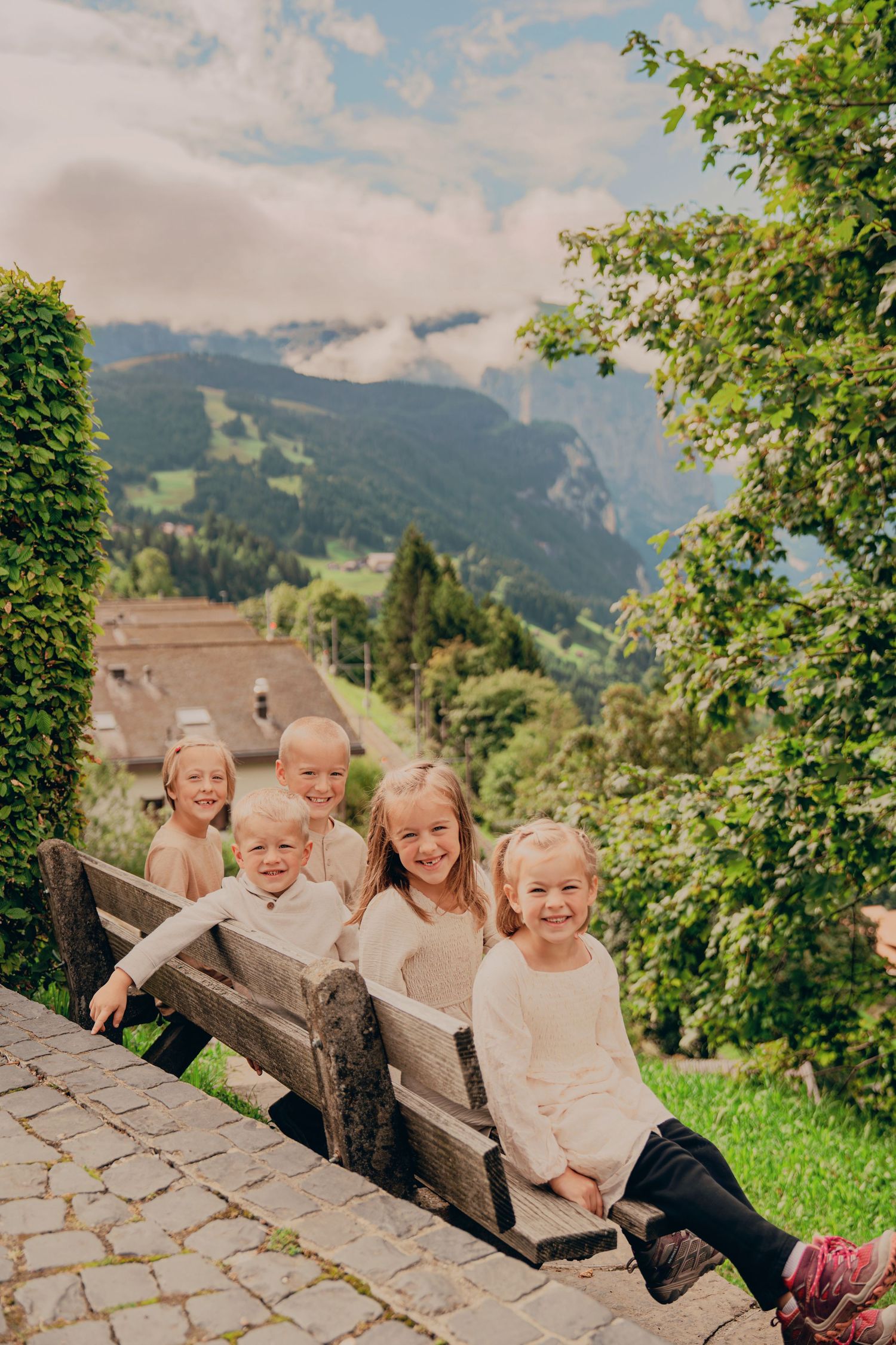 Five cheerful children sitting on a wooden bench with stunning Swiss Alps in background.