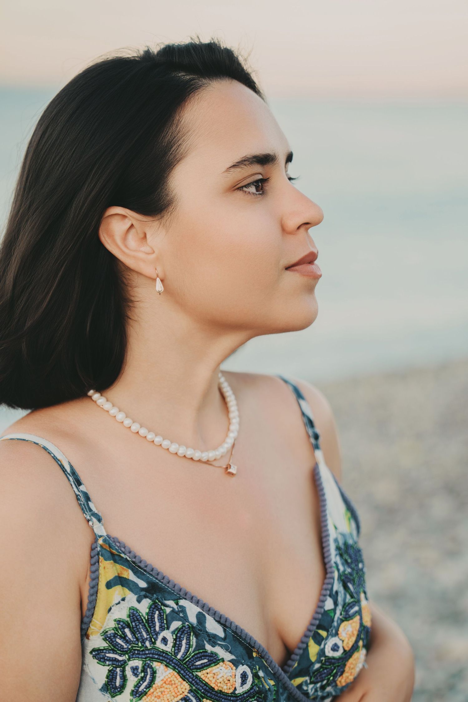 Close-up side profile of young brunette woman wearing pearl necklace and earrings in floral dress at beach.