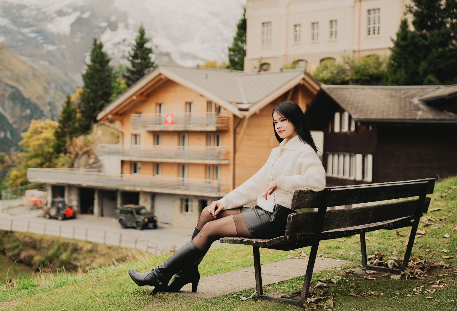 Young Asian woman in white sweater and black boots sits on a bench with Alpine village and mountains backdrop.