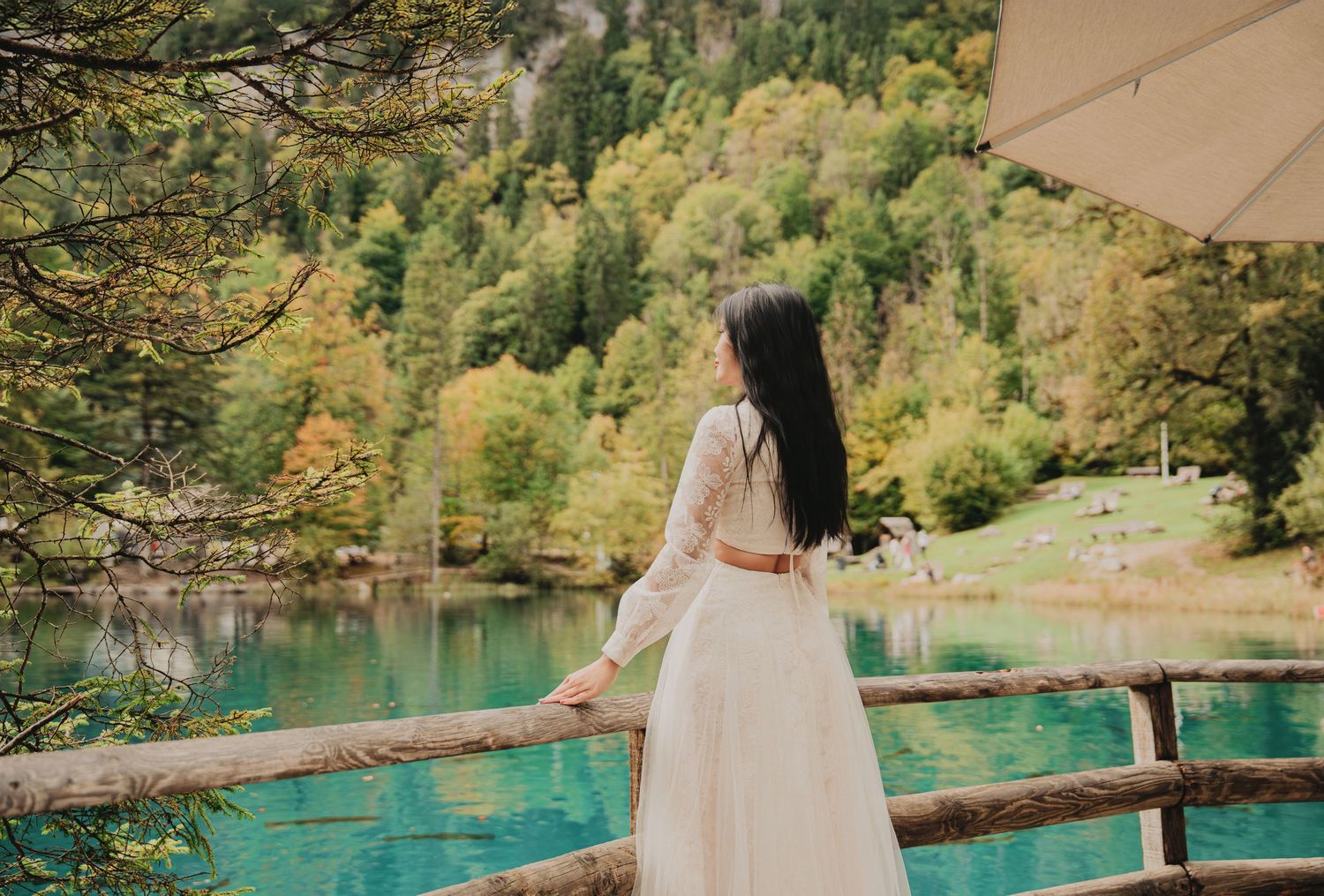 Woman in white dress leans on rustic wooden fence overlooking serene turquoise mountain lake.
