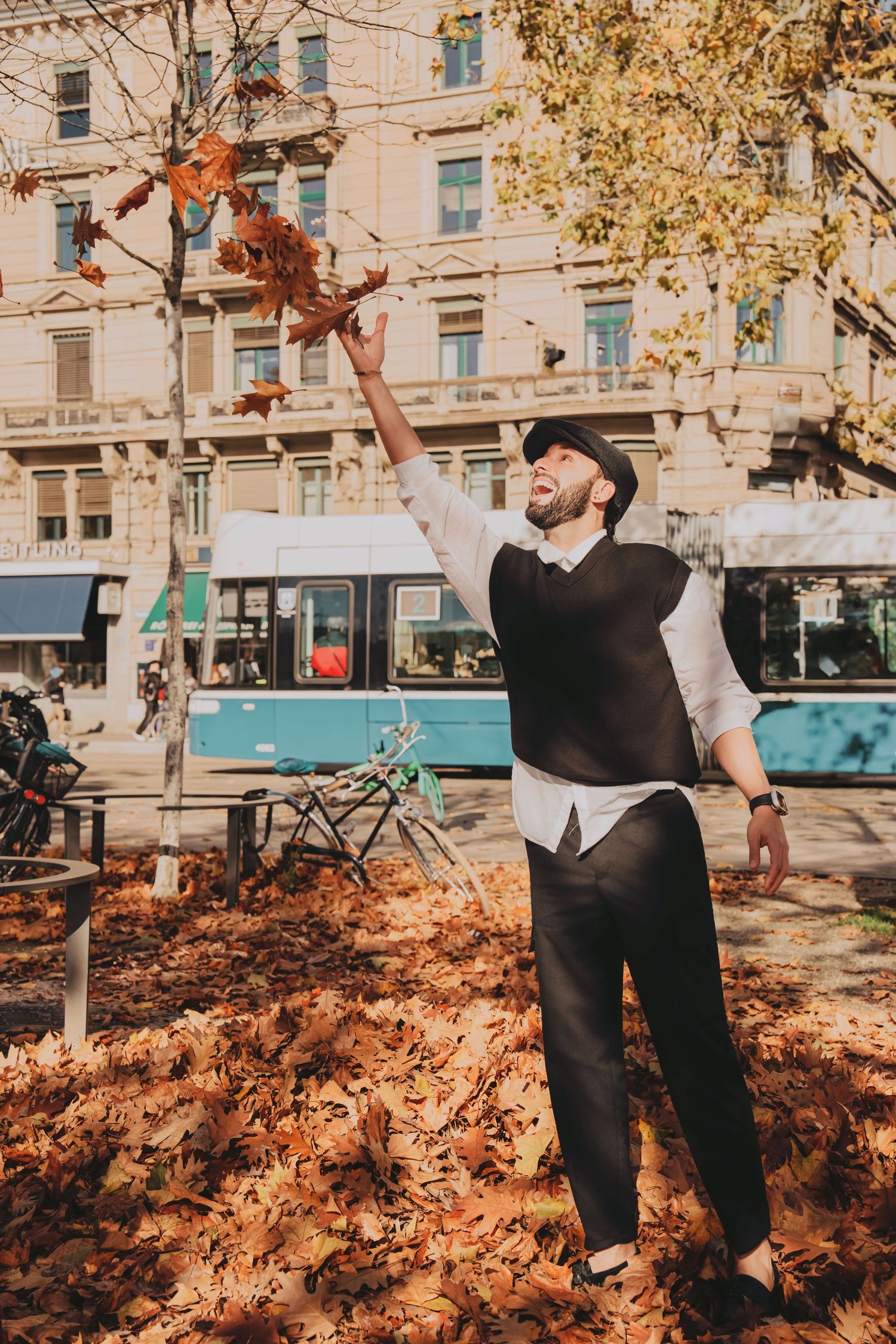 Joyful man in flat cap tossing autumn leaves in sunny urban square with tram behind.