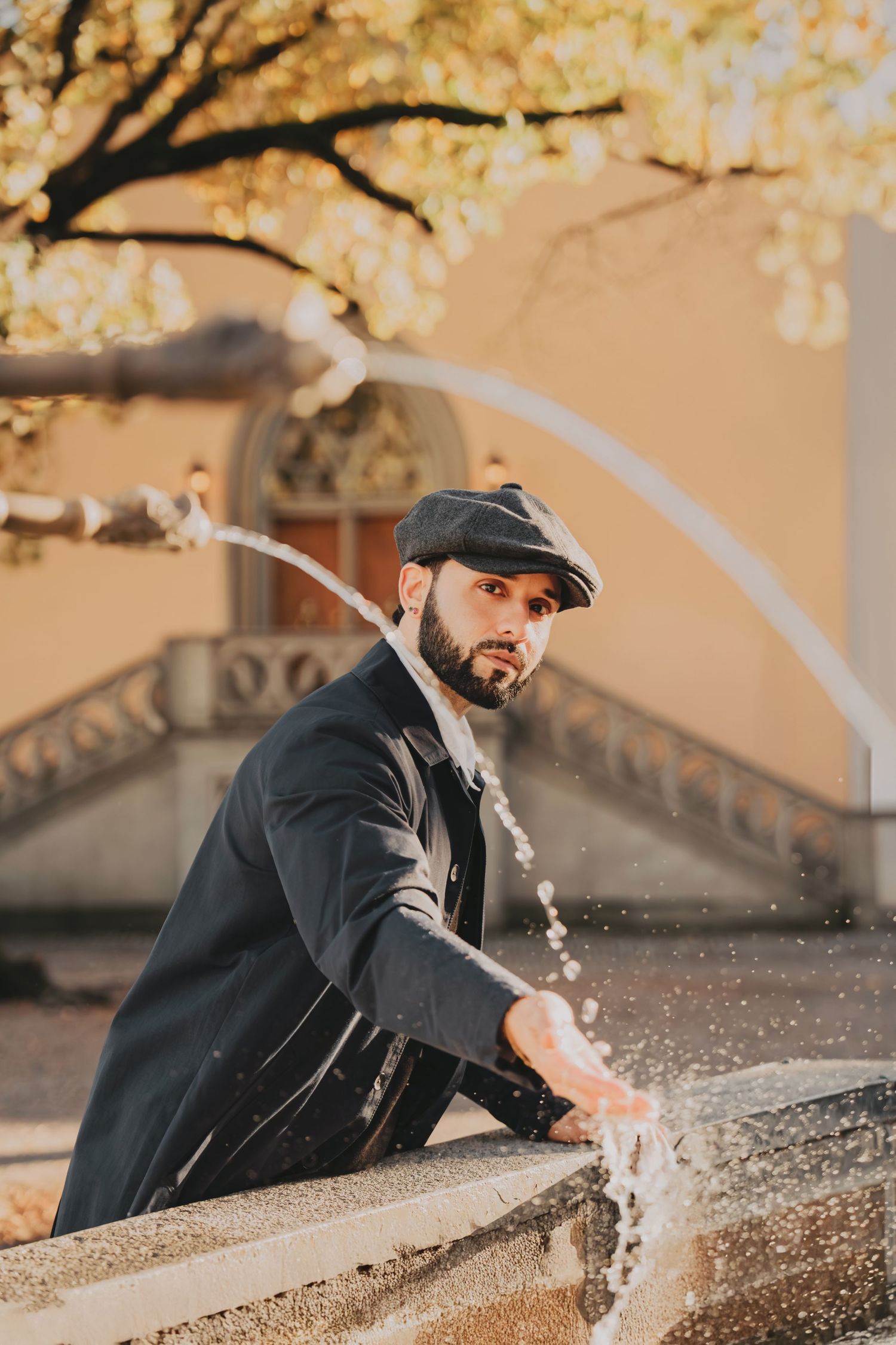 Stylish bearded man in flat cap splashing water at an ornate stone fountain in autumn sunlight.
