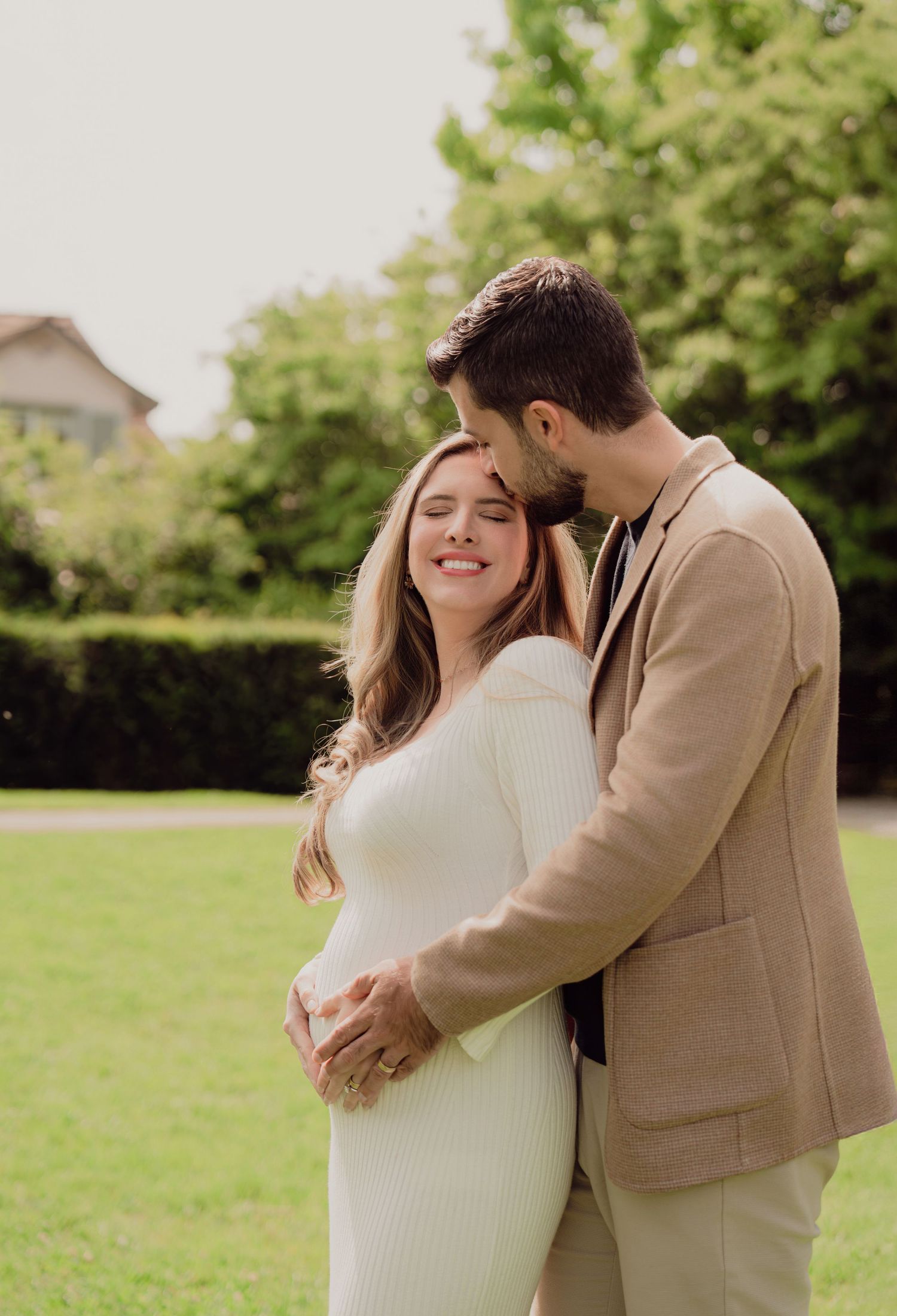 Man tenderly kissing pregnant partner's head while embracing her baby bump in a green park.