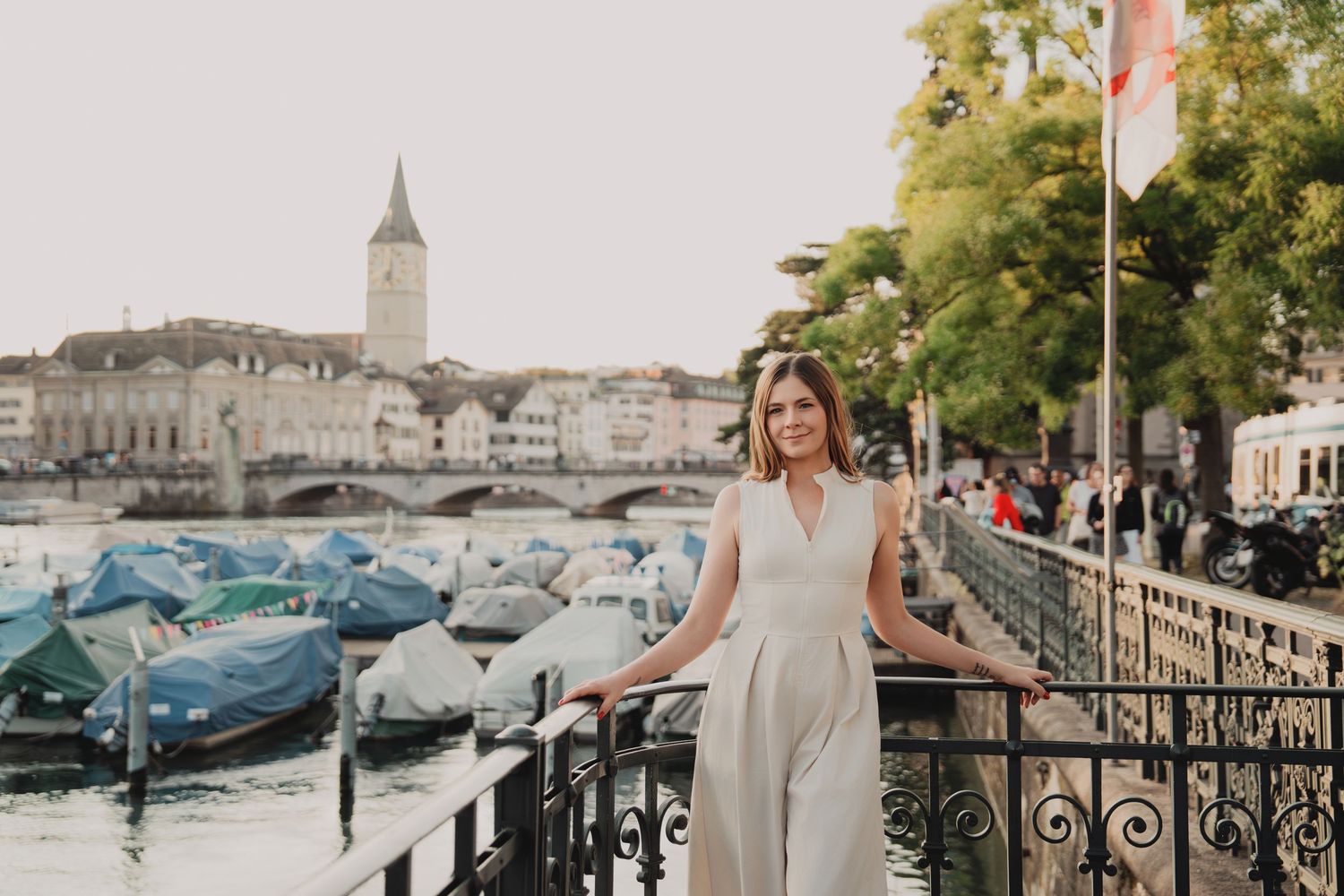 Woman in white jumpsuit posing by iron railing with Zurich old town and river in background.
