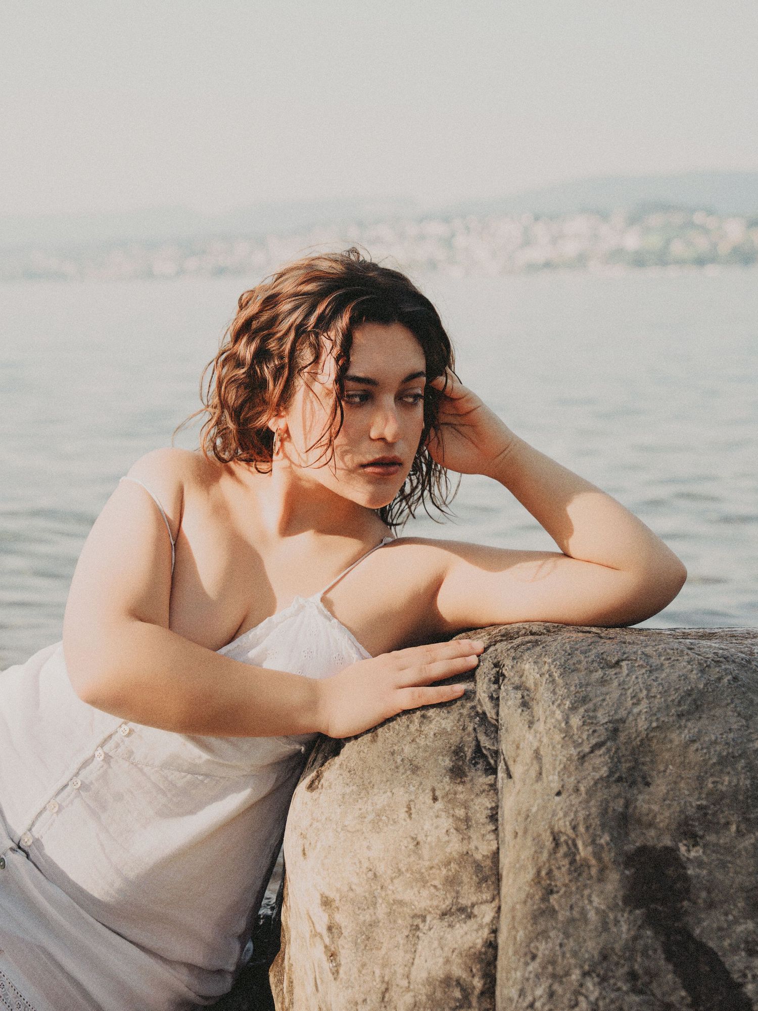 Woman with wet hair in white dress resting on rock by the seaside.