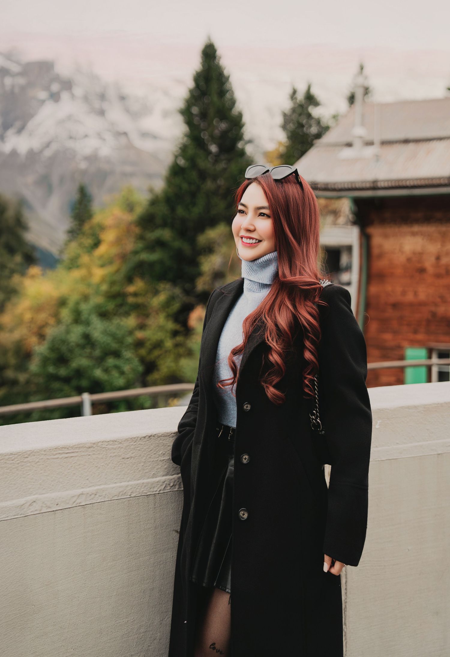 Smiling woman with long red wavy hair wearing black long coat and turtleneck with mountain backdrop.