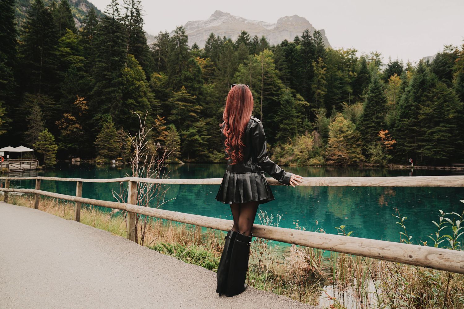 Back view of red-haired woman in black leather outfit standing by turquoise mountain lake and forest.