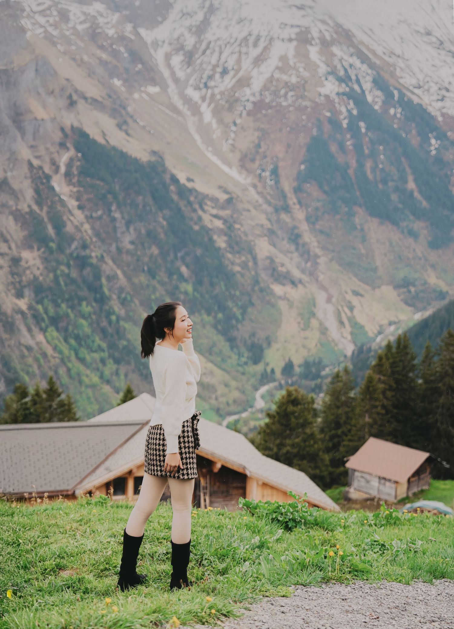 Smiling woman in white sweater and plaid skirt poses on lush green alpine meadow with snowy mountains.